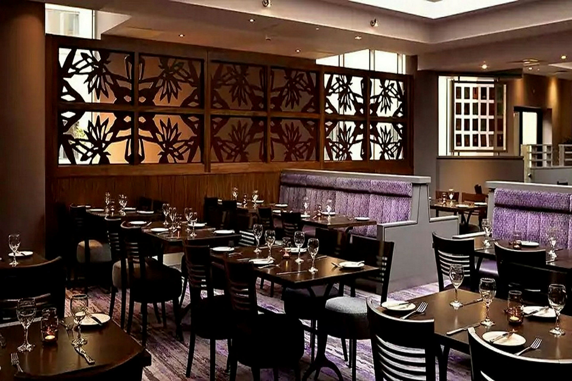 En stilfull restaurang med bord och stolar uppställda, en dekorativ trävägg med blommönster och lila detaljer på sittplatser. (AI-genererad text)