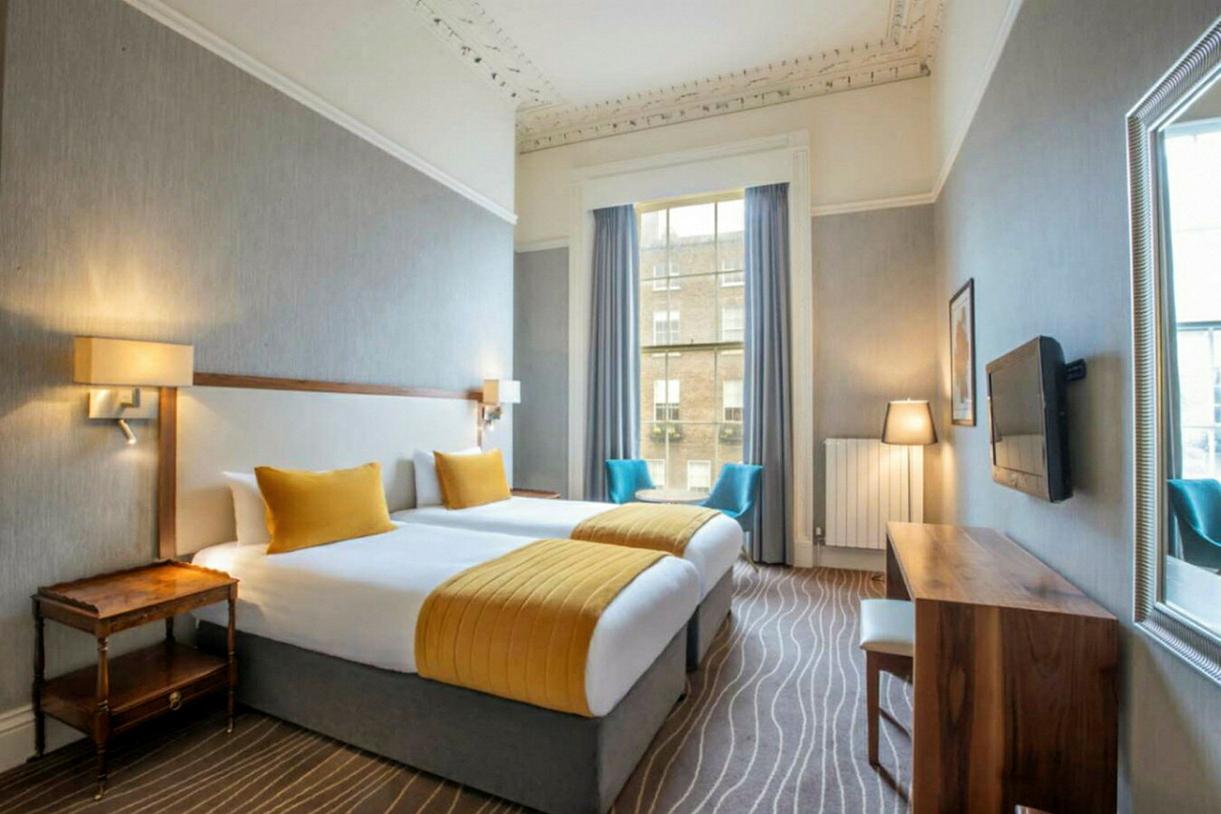 Ett hotellrum med två separata sängar, guldfärgade sängöverkast och kuddar, ett träskrivbord med stol, TV på väggen, och en stor fönsteröppning med blå gardiner och utsikt utåt. (AI-genererad text)