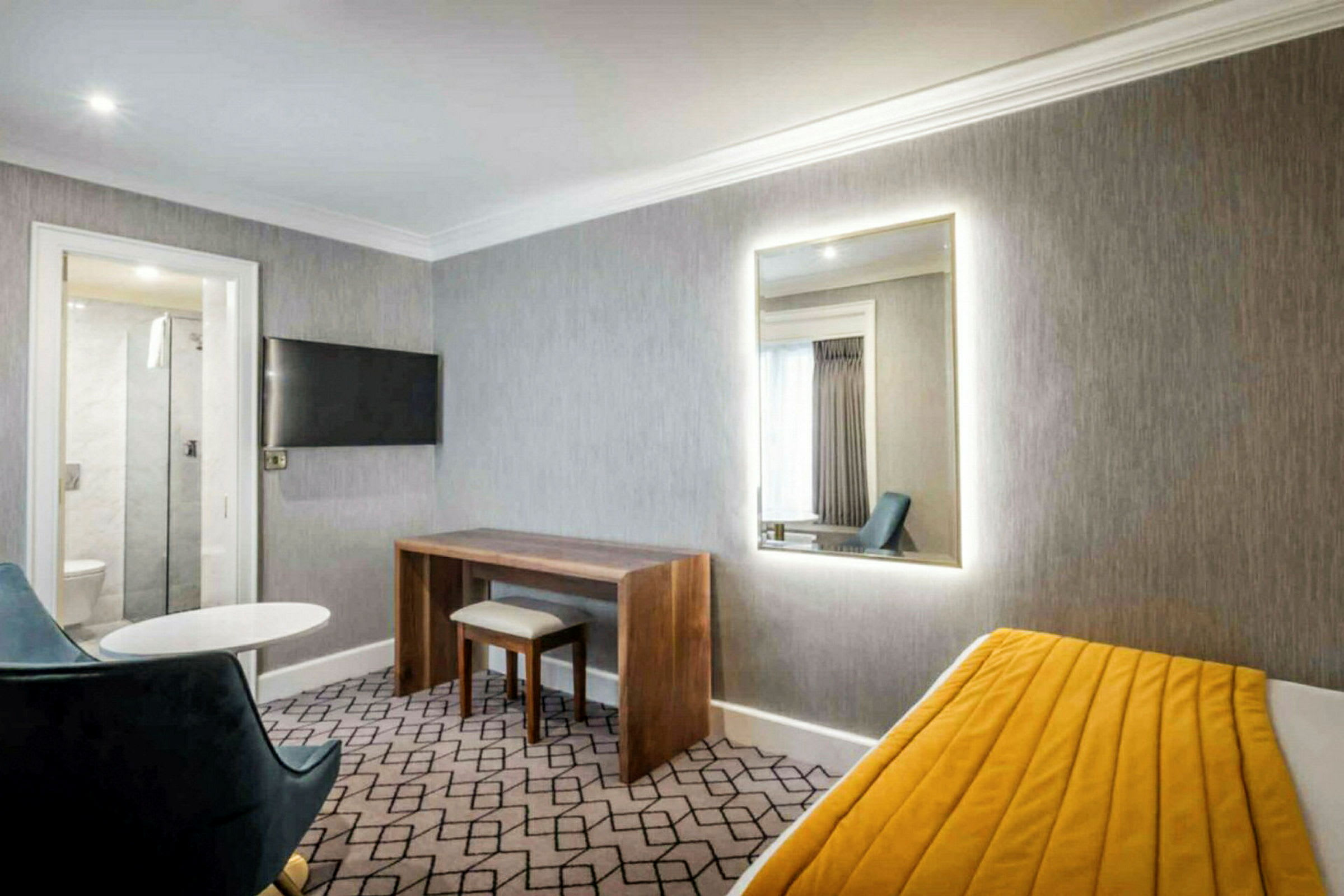Ett hotellrum med grå väggar, en spegel, ett skrivbord, en stol och en säng med gul filt. (AI-genererad text)