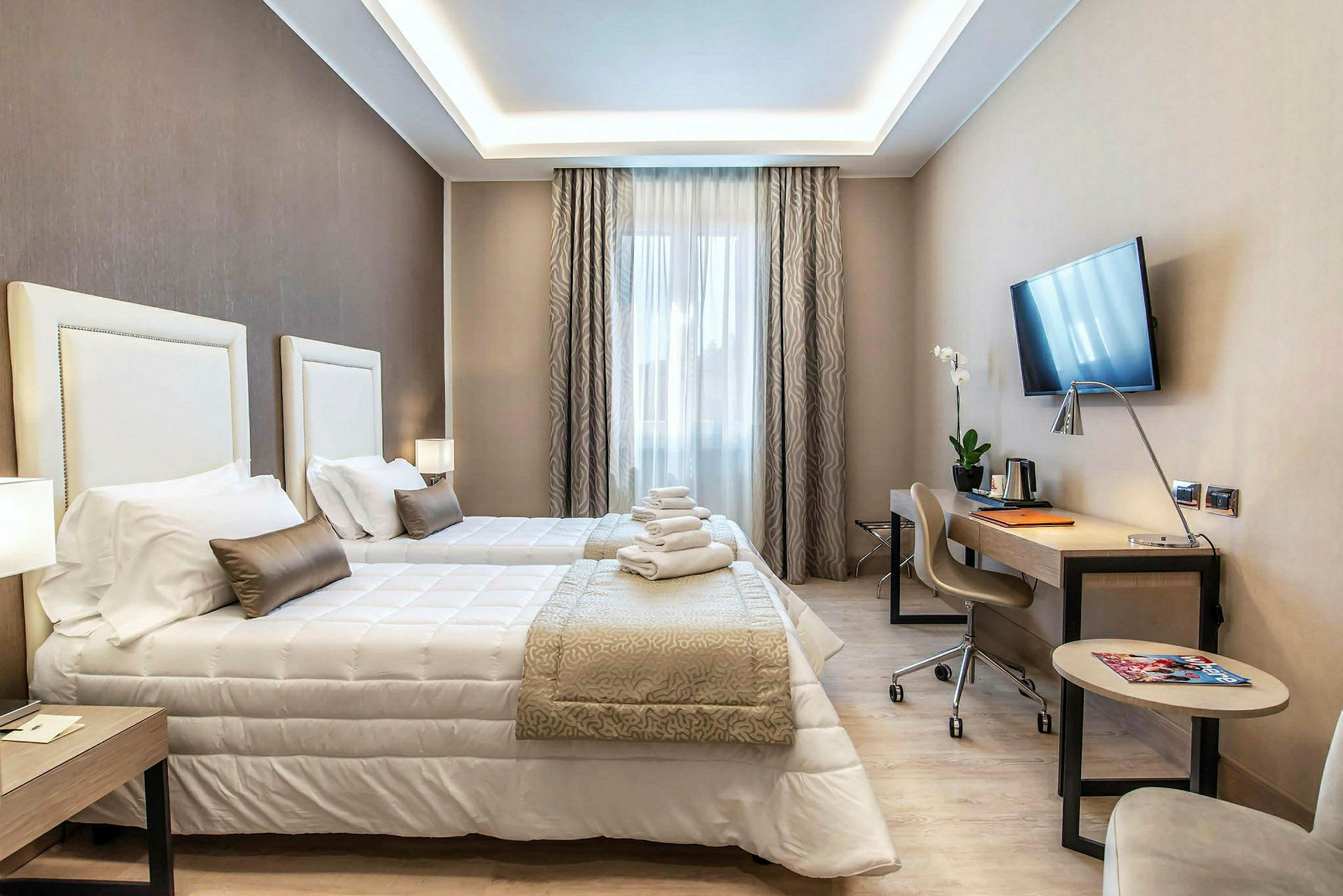 Ett hotellrum med två enkelsängar, ett skrivbord, en väggmonterad TV och en stol. Rummet är inrett i ljusa färger med snygga gardiner. (AI-genererad text)