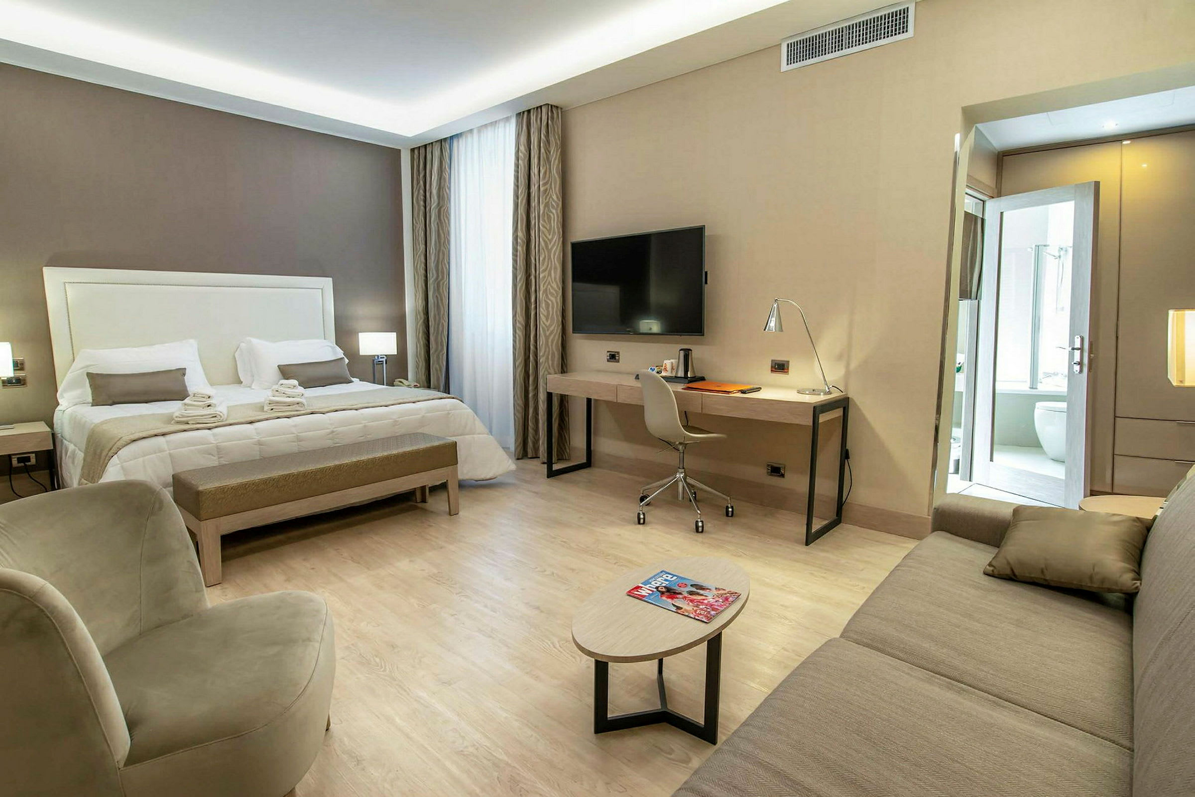 Ett hotellrum med stor säng, skrivbord, fåtölj, soffa och en liten bord, ljus och modern inredning. (AI-genererad text)