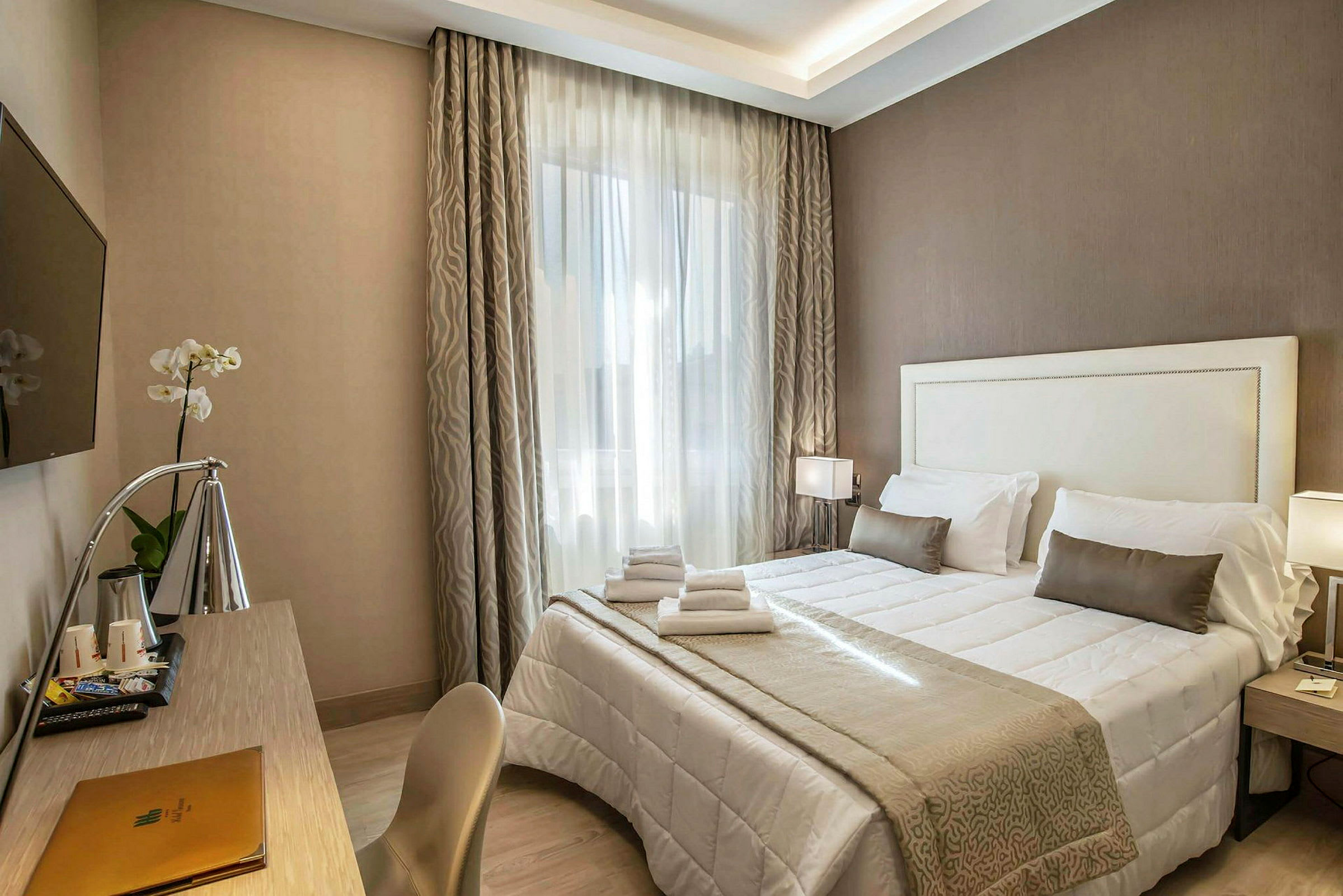 Ett hotellrum med en dubbelsäng täckt med vita och beige sängkläder, ett skrivbord med en lampa och en väggmonterad TV. (AI-genererad text)