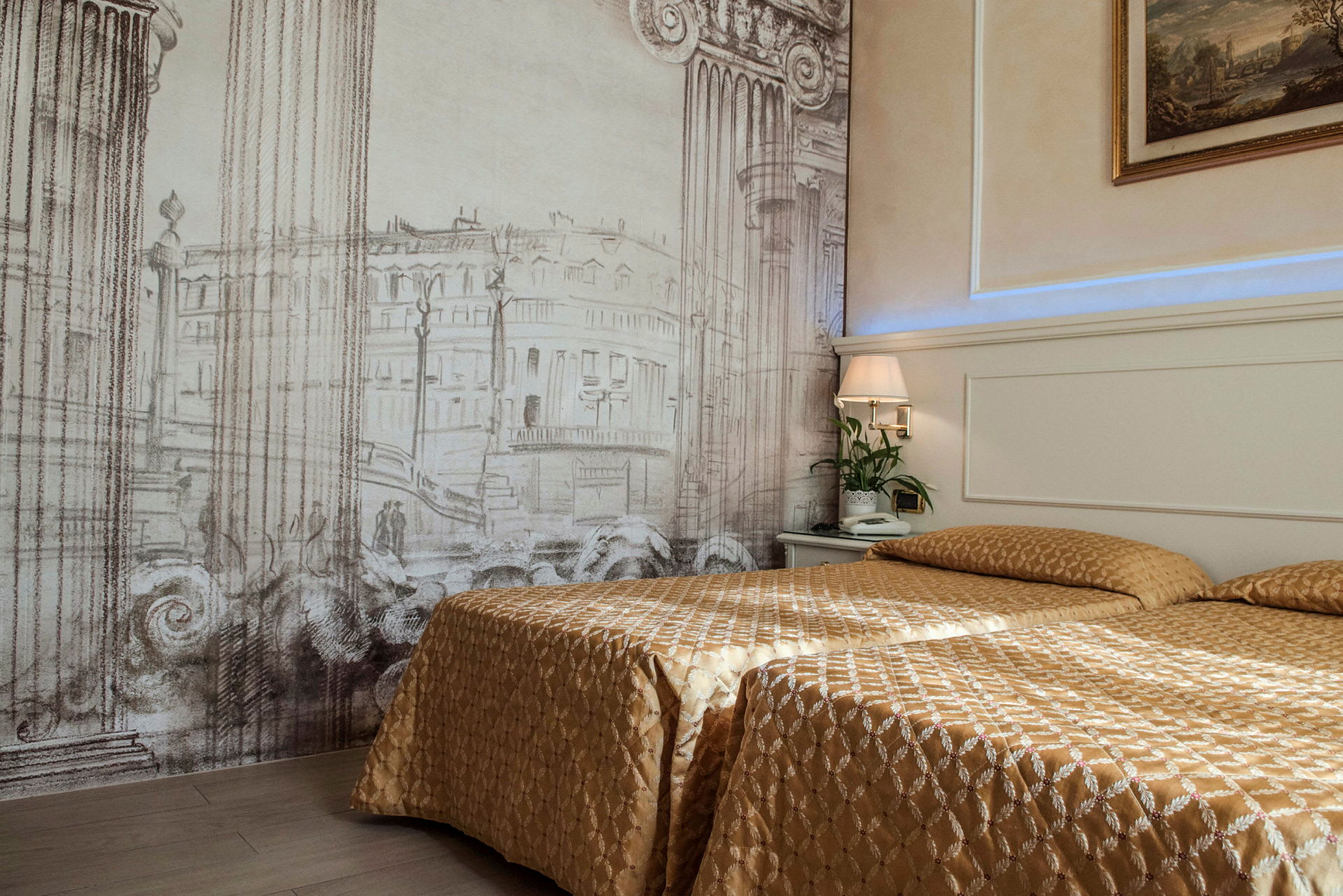 Ett hotellrum med två sängar täckta av guldfärgade överkast. Bakgrundsväggen har ett konstverk med arkitektoniska pelare och byggnader. En nattlampa och en växt står på nattduksbordet. (AI-genererad text)