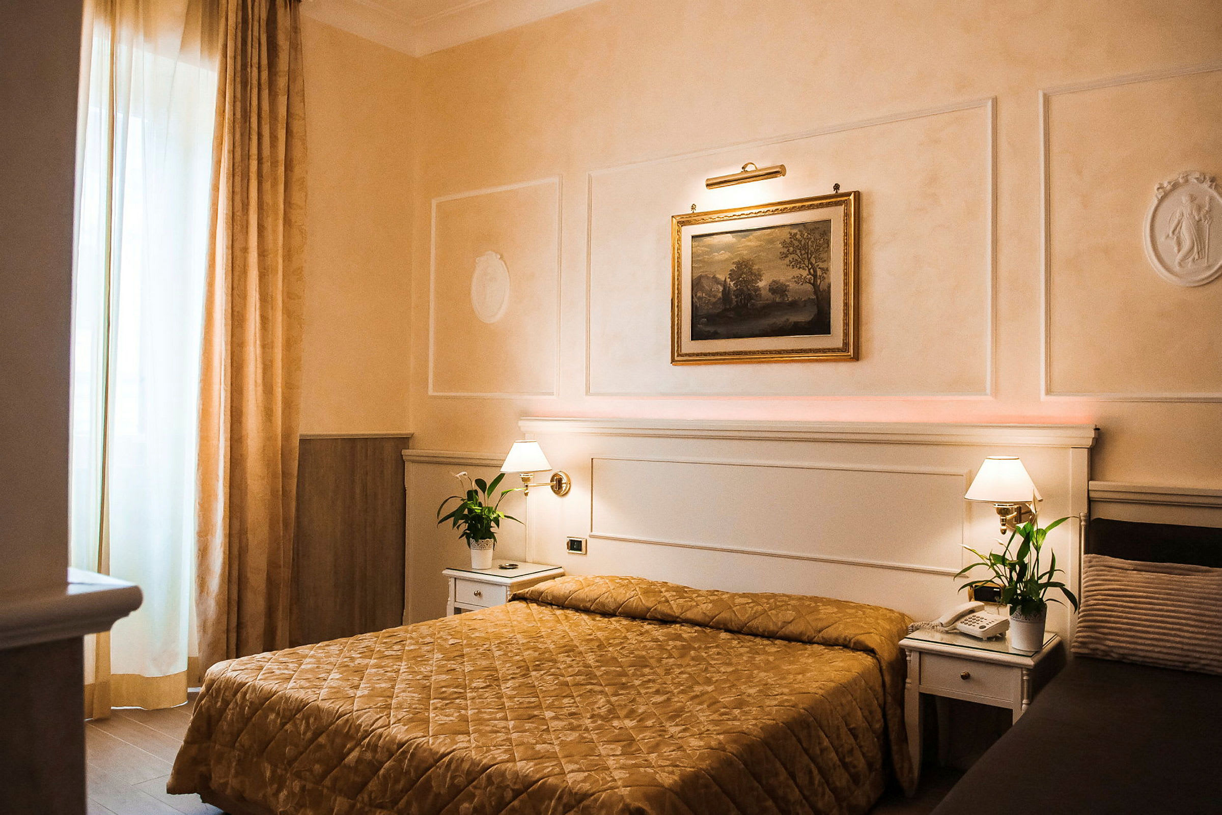 Ett hotellrum med en säng täckt av ett guldfärgat överkast, två nattduksbord med lampor och gröna växter, samt en tavla på väggen ovanför sängen. (AI-genererad text)