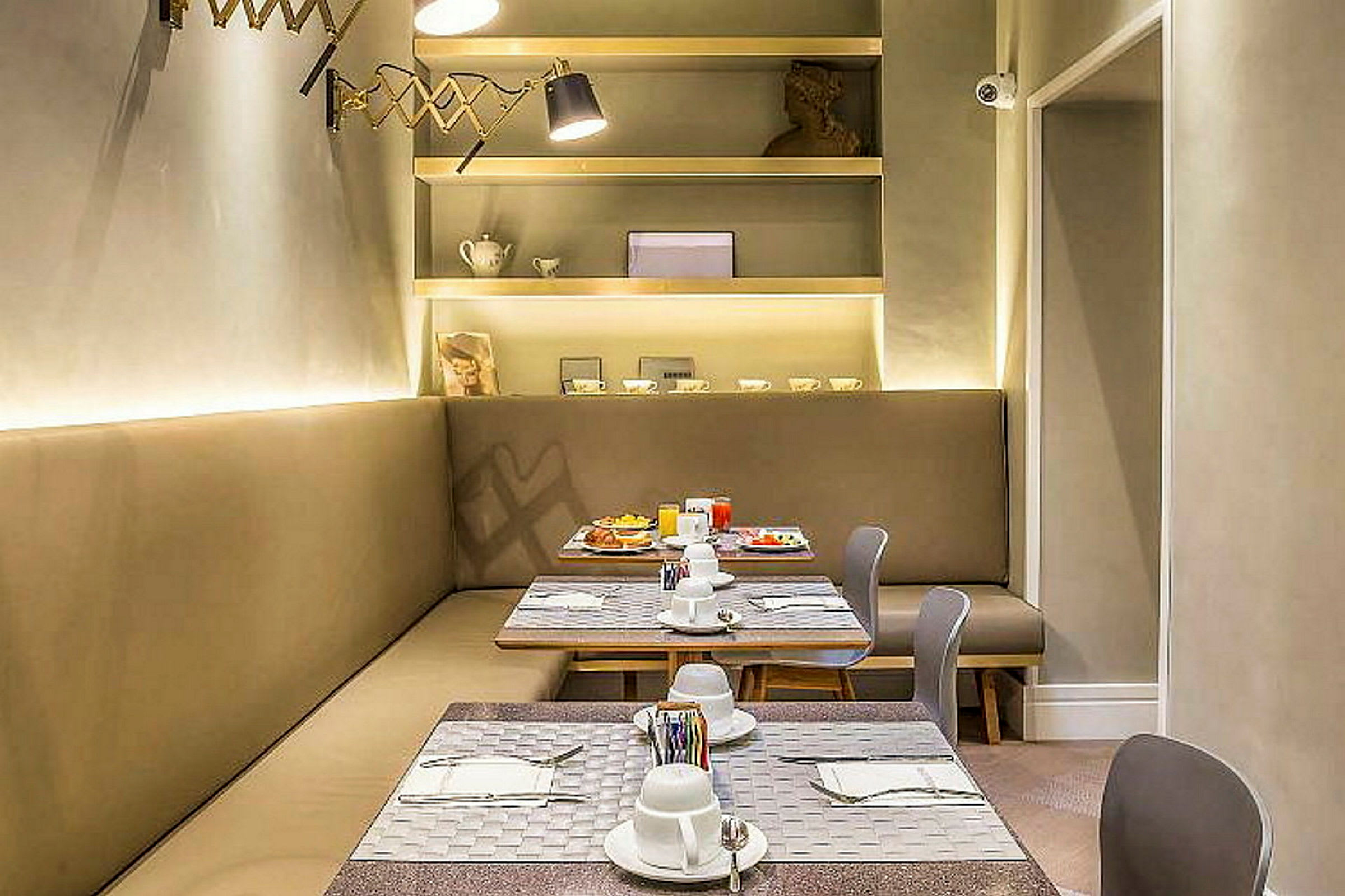 Ett modernt möblerat restaurangområde med bord, stolar och en sittbänk. Bordet är dukat med porslin och bestick. På väggen finns hyllor med dekorativa föremål. (AI-genererad text)