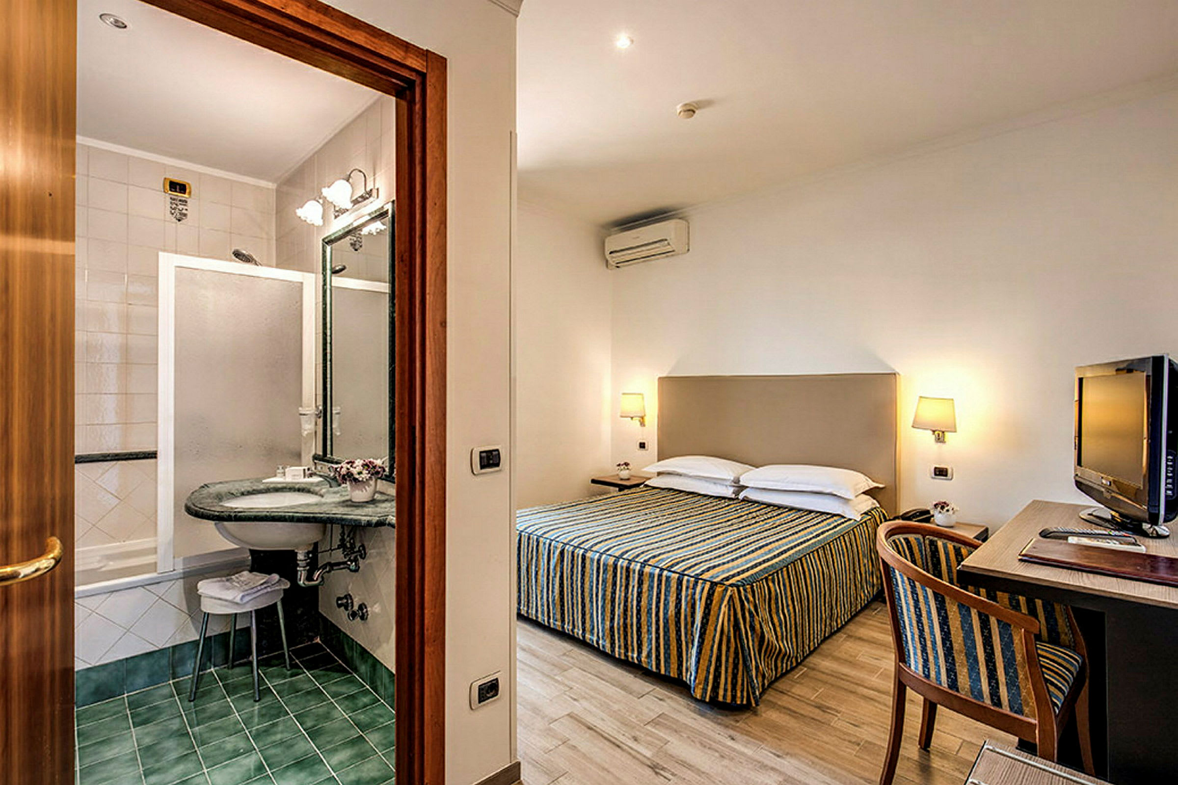 Ett hotellrum med dubbelsäng, ett skrivbord med stol och en TV. Ett angränsande badrum med dusch och handfat. (AI-genererad text)