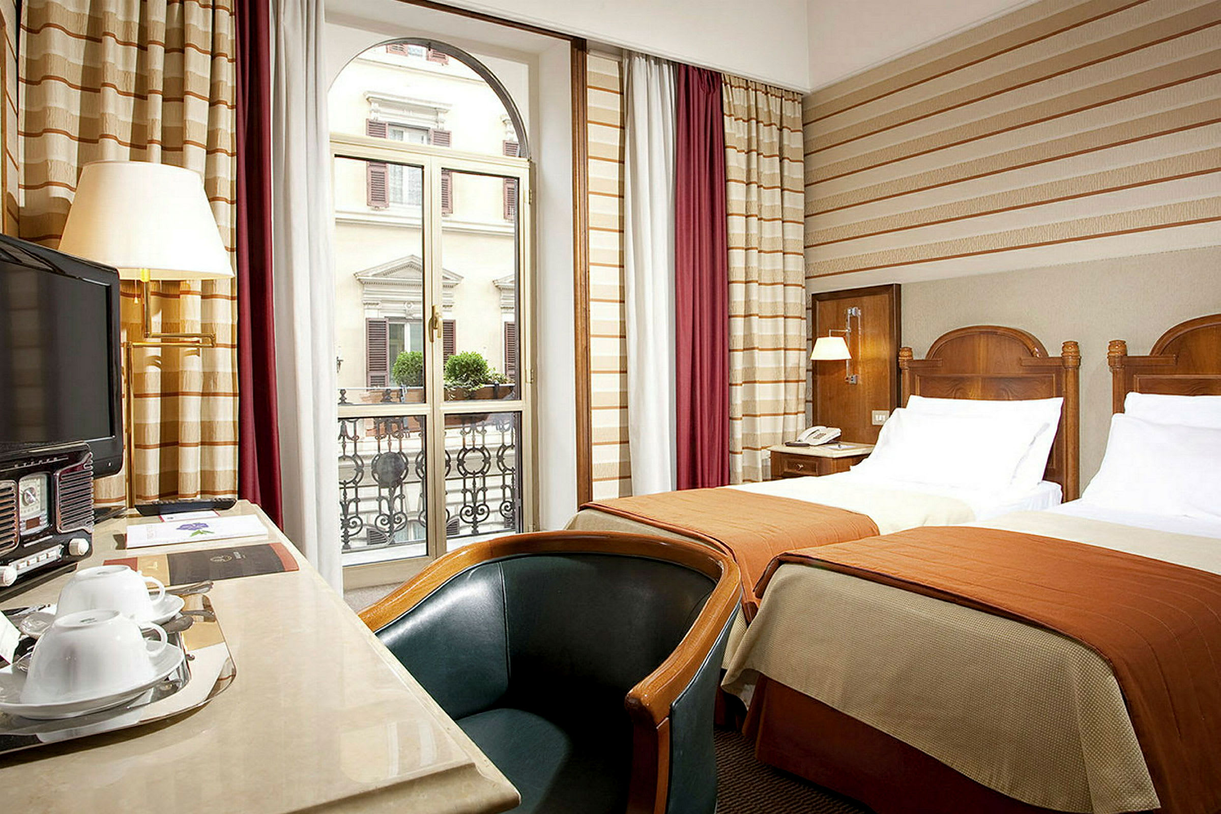 Hotellrum med två enkelsängar och sittmöbler, utsikt mot en byggnad med balkong. (AI-genererad text)