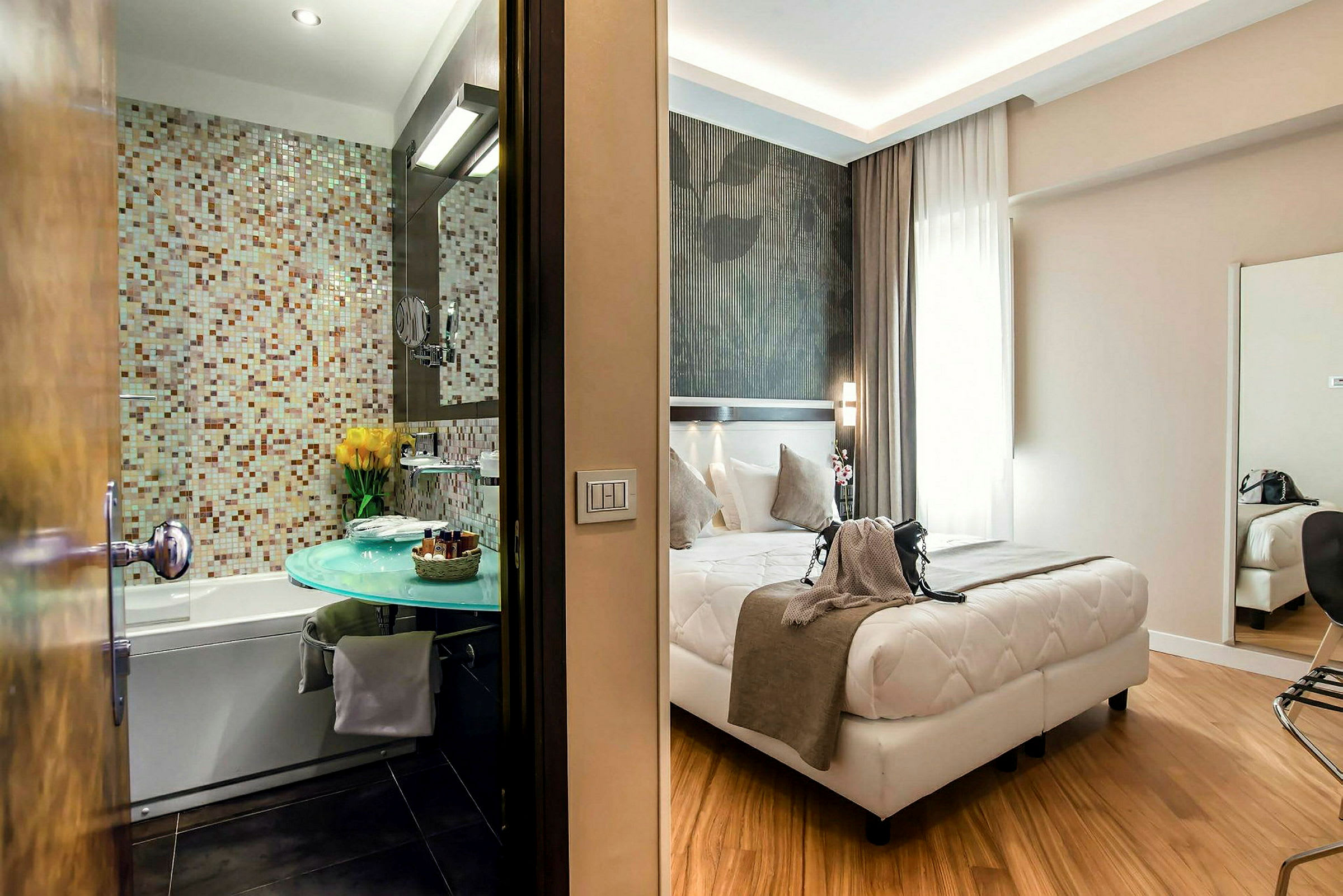 Ett hotellrum med en säng framför ett stort fönster, samt ett badrum med mosaikvägg och ett modernt tvättställ. (AI-genererad text)