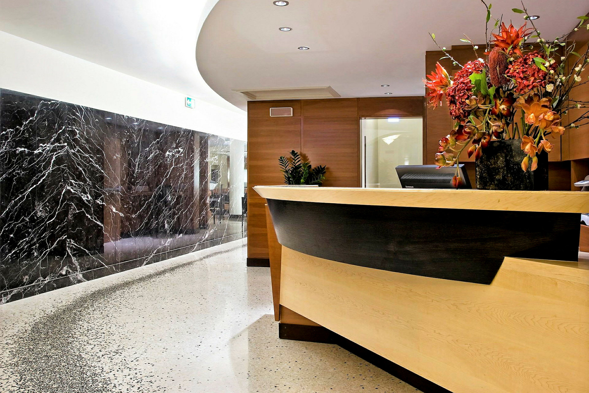 En receptionsdisk på ett hotell, dekorerad med en stor vas med blommor och omgiven av marmordetaljer och elegant belysning. (AI-genererad text)