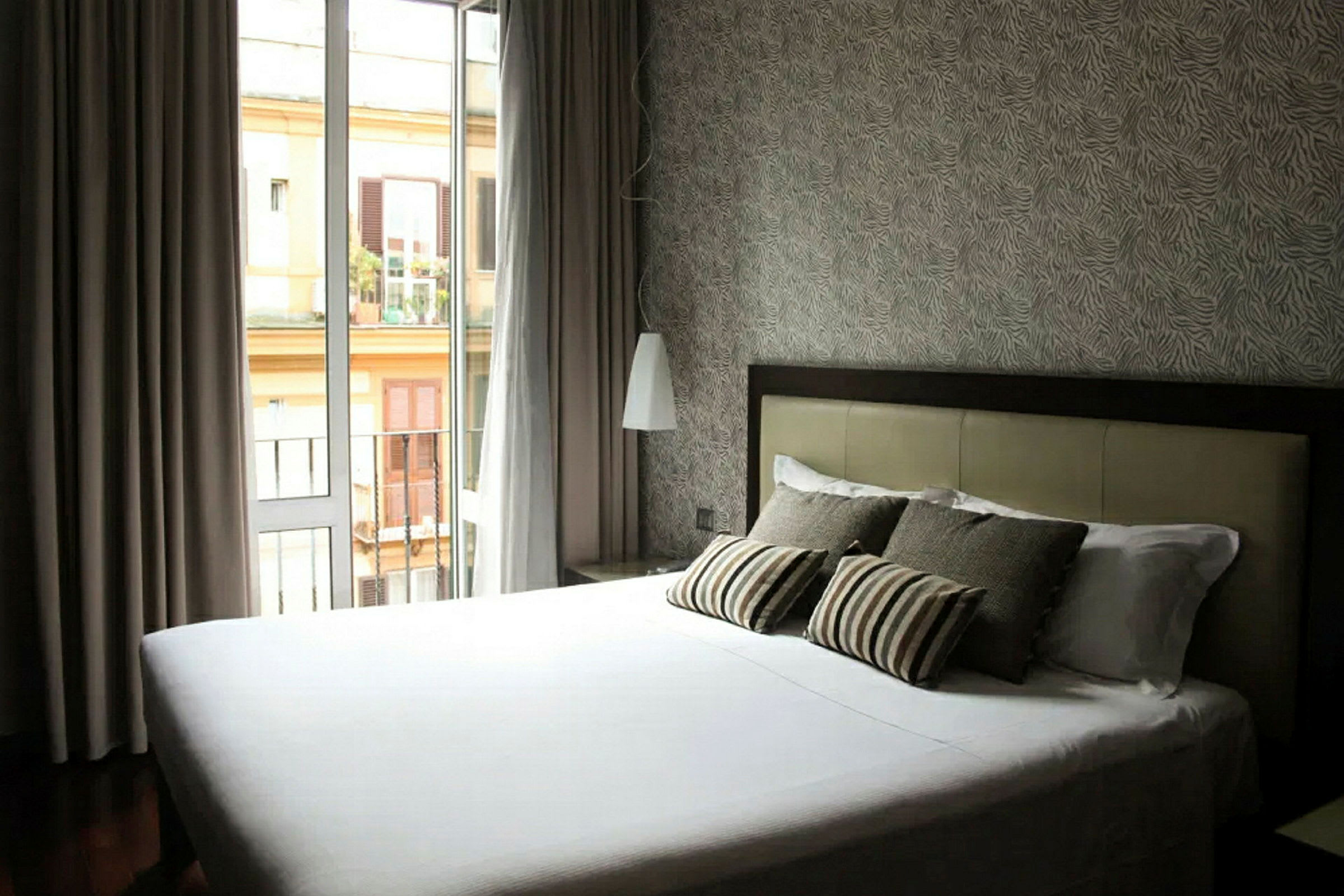 Ett hotellrum med en säng klädd i vita lakan och flera kuddar, en närliggande balkongdörr som släpper in naturligt ljus, och detaljerad tapet på väggen. (AI-genererad text)