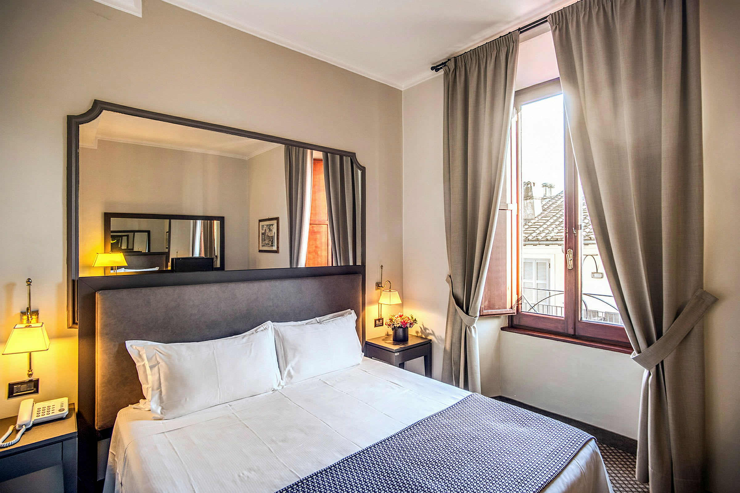 Hotellrum med dubbelsäng, sängbord, spegel och fönster med beige gardiner. (AI-genererad text)
