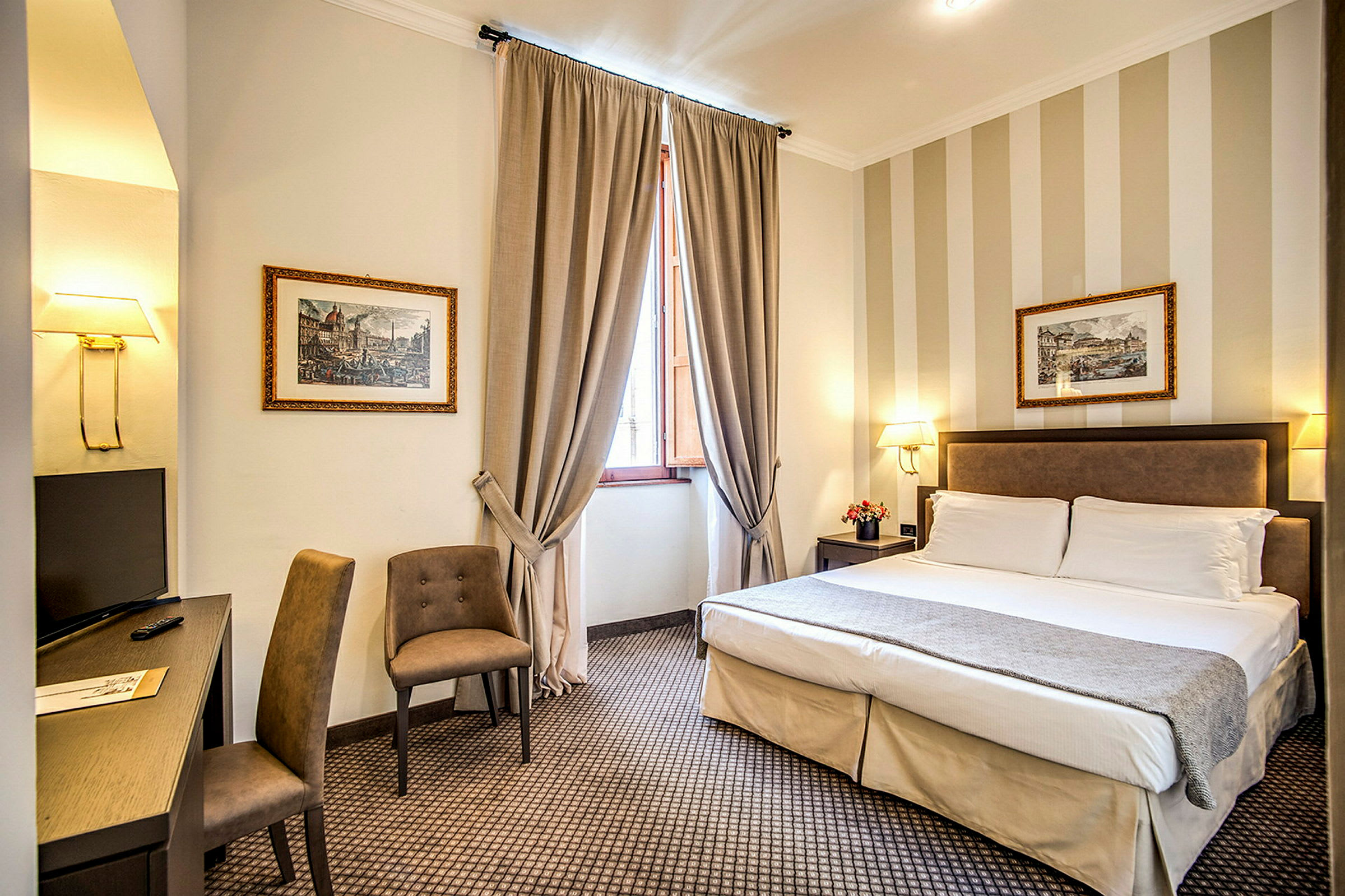 Ett hotellrum med en dubbelsäng, två stolar, skrivbord och en TV. Rummet har gardiner vid fönstret och dekorativa tavlor på väggen. (AI-genererad text)