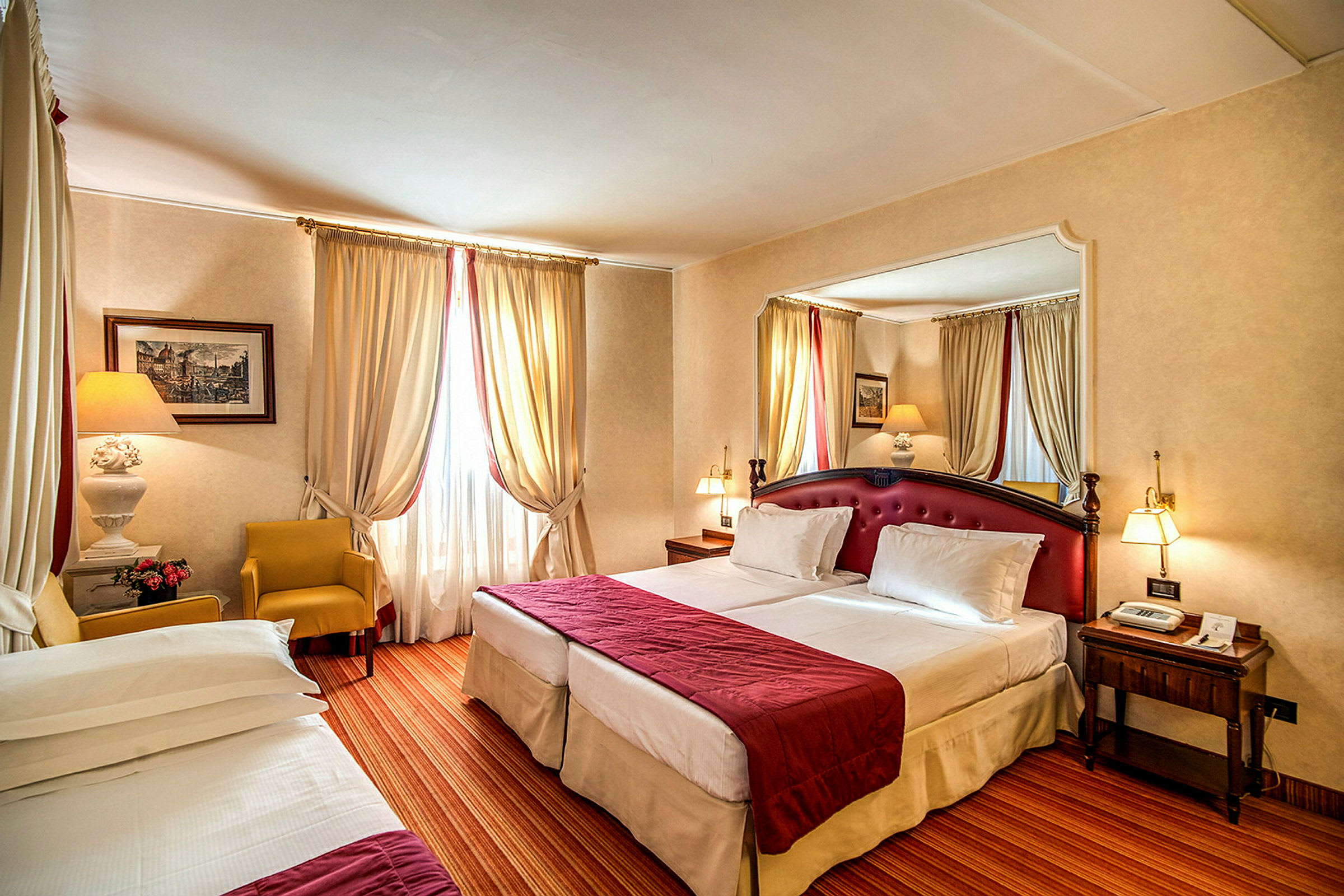 Ett hotellrum med två enkelsängar med röda överkast och vita sängkläder, en gul fåtölj, ett sidobord och gardiner vid ett fönster. (AI-genererad text)