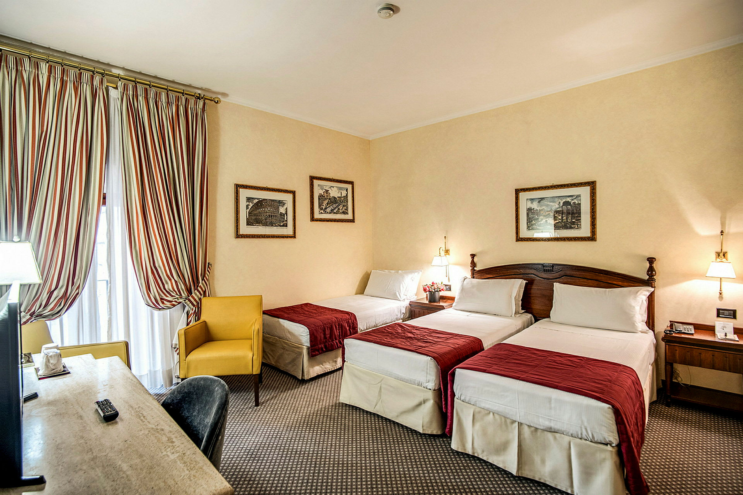 Hotellrum med tre enkelsängar och röd sängklädsel. En gul fåtölj vid fönstret samt ett skrivbord med TV. (AI-genererad text)
