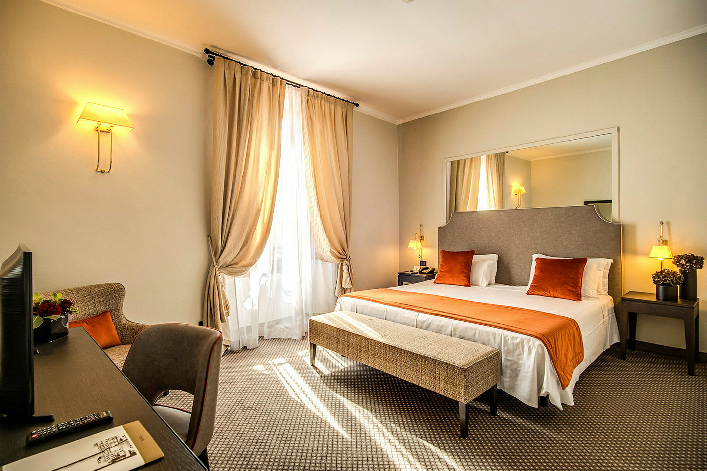 Ett hotellrum med en dubbelsäng, nattduksbord och en bänk vid sängens fotände. Gardinen draperar fönstret och en skrivbord med en stol står bredvid. (AI-genererad text)