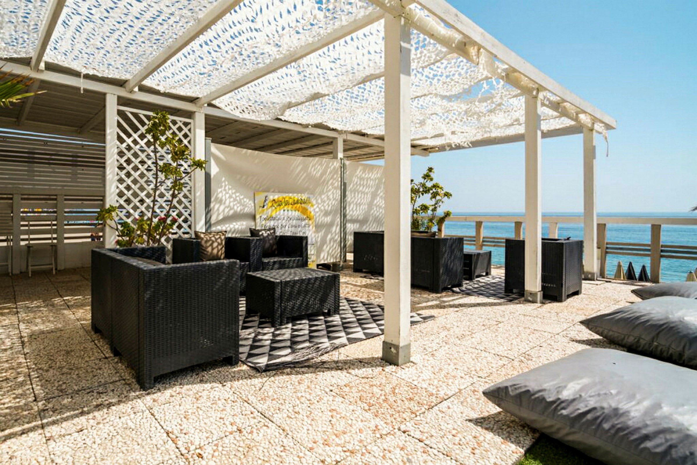 Utomhus sittplats med svart rottingmöbler under en vit pergola med utsikt över havet. (AI-genererad text)