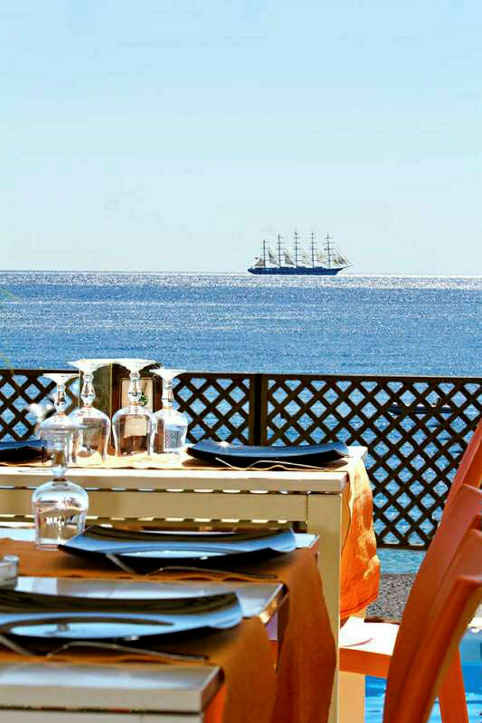 Ett dukat bord på en terrass med utsikt över havet och ett segelfartyg i fjärran. (AI-genererad text)