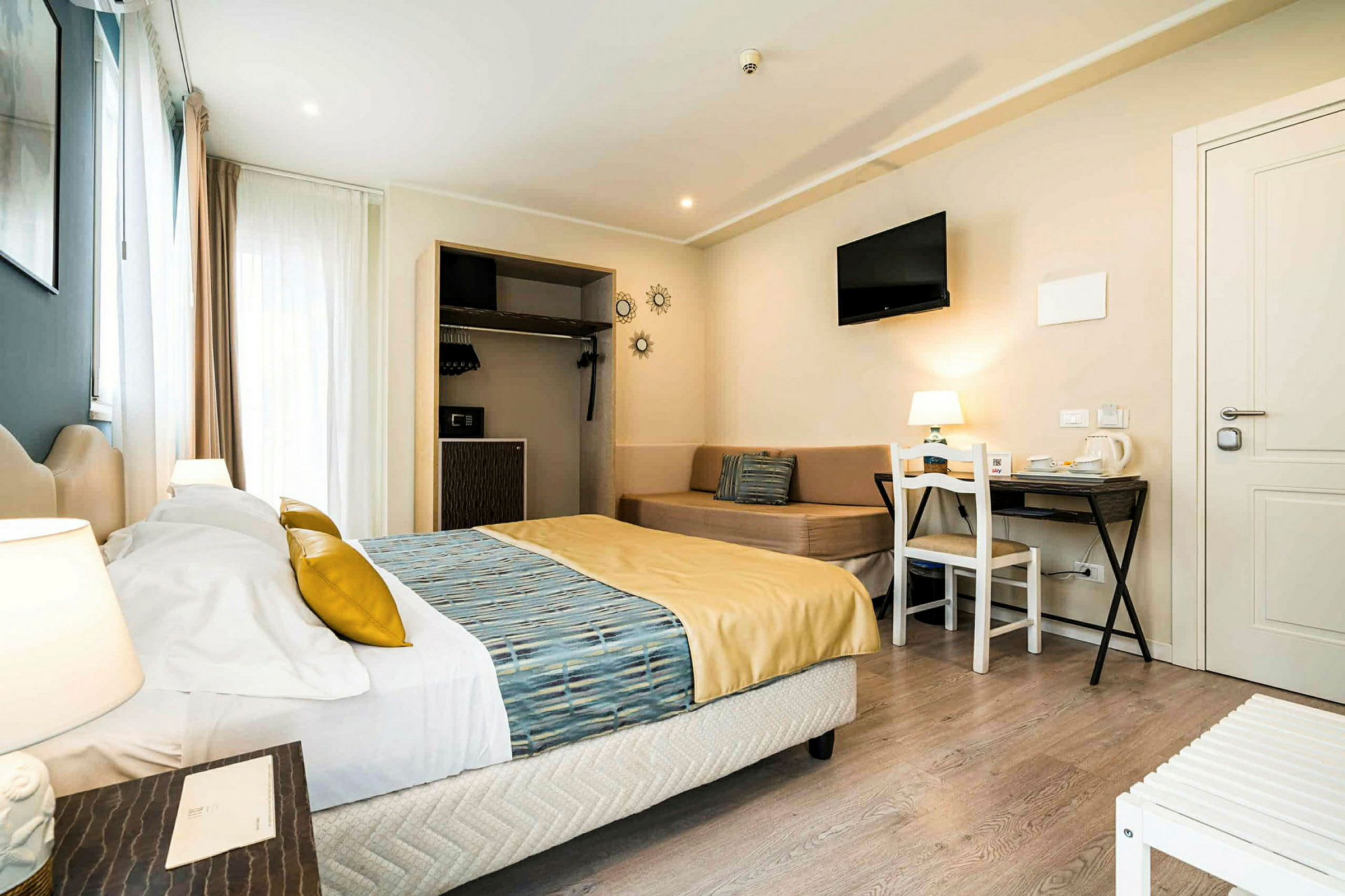 Ett hotellrum med en dubbelsäng bredvid ett nattduksbord, en soffa, ett skrivbord och ett väggmonterat tv. (AI-genererad text)
