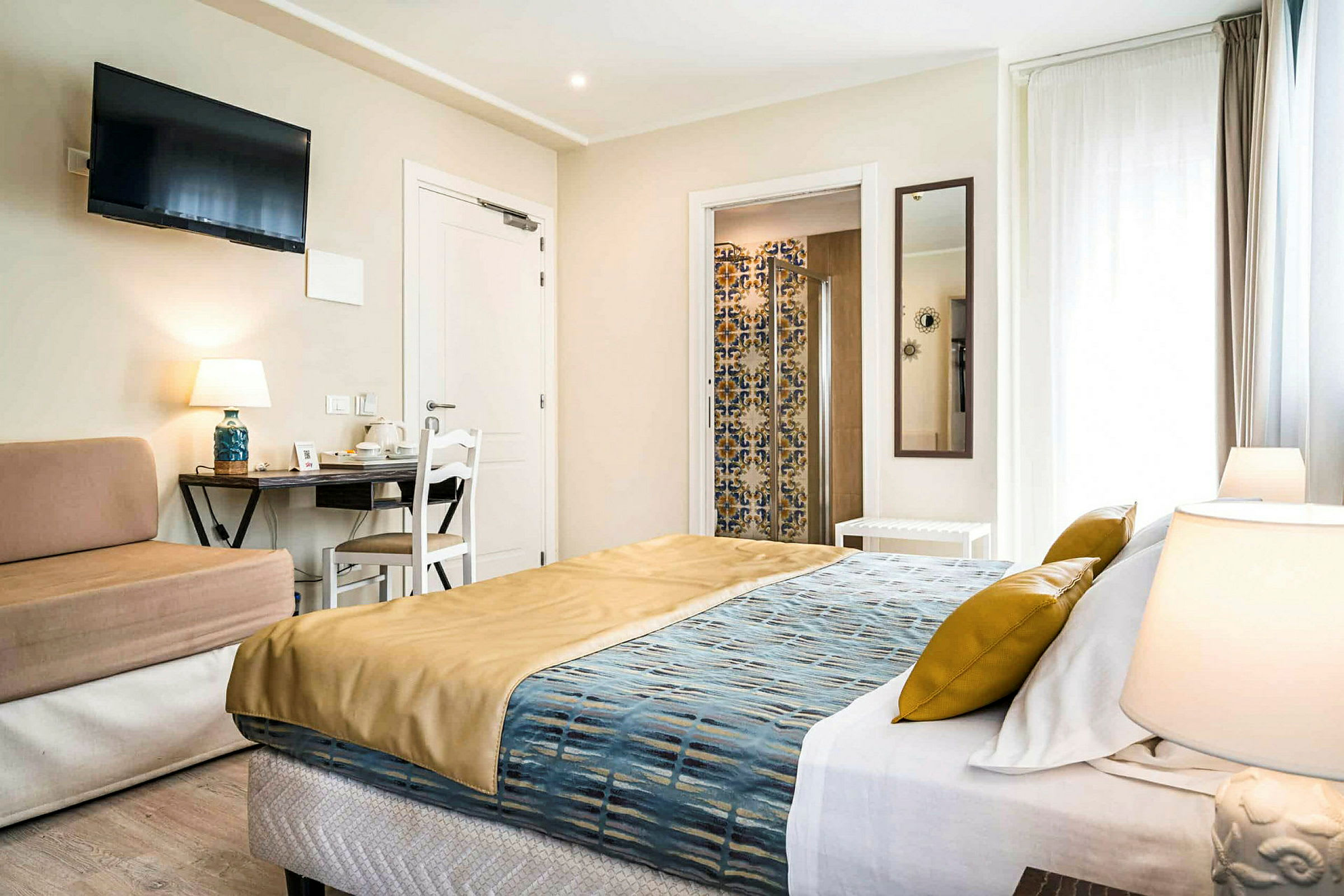 Ett hotellrum med en stor säng, en soffa, ett skrivbord och en TV på väggen. Ett litet badrum med en dusch syns i bakgrunden. (AI-genererad text)