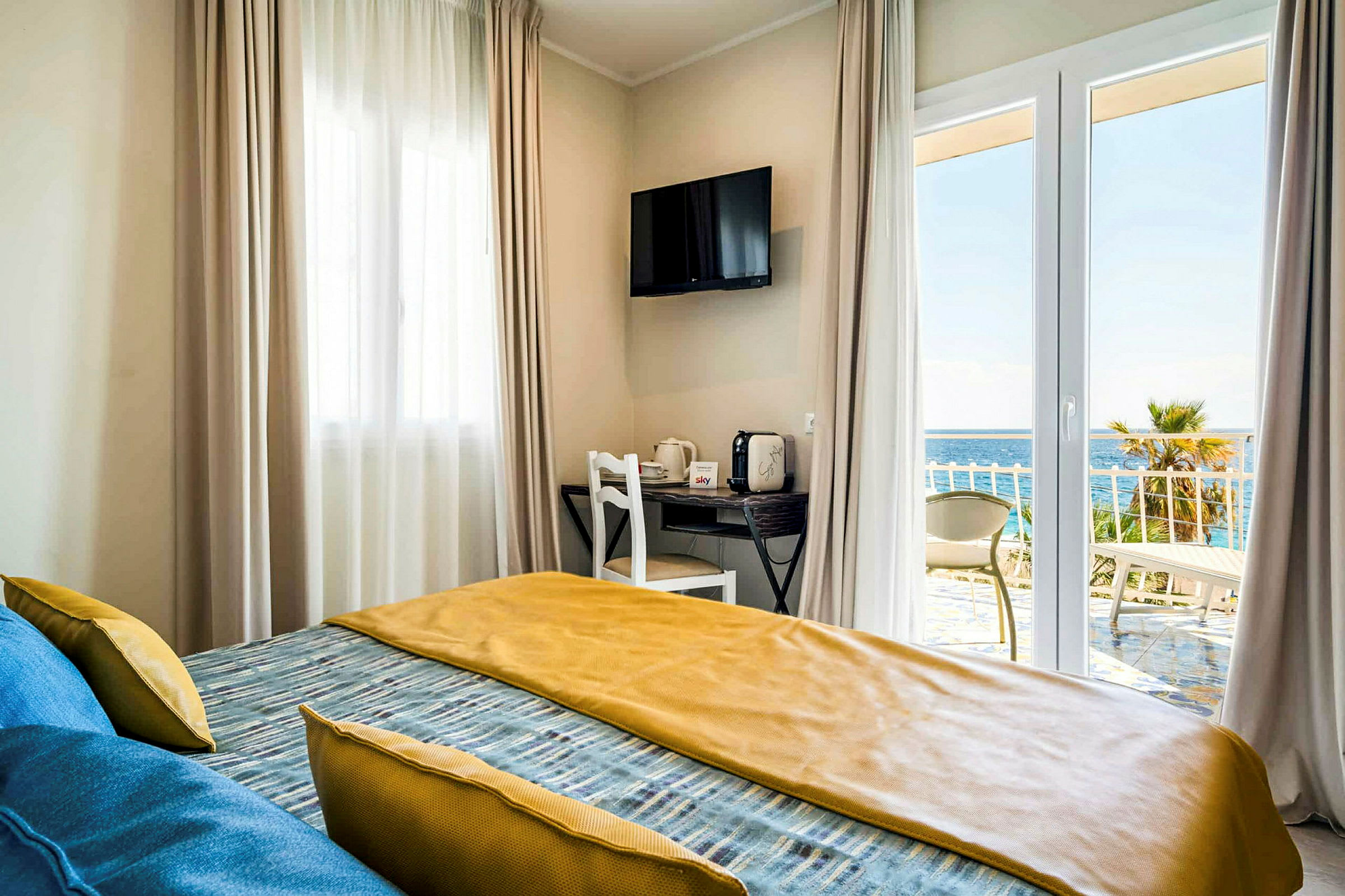 Ett hotellrum med en säng, fönster med gardiner, en TV på väggen och utsikt över havet från balkongen. (AI-genererad text)