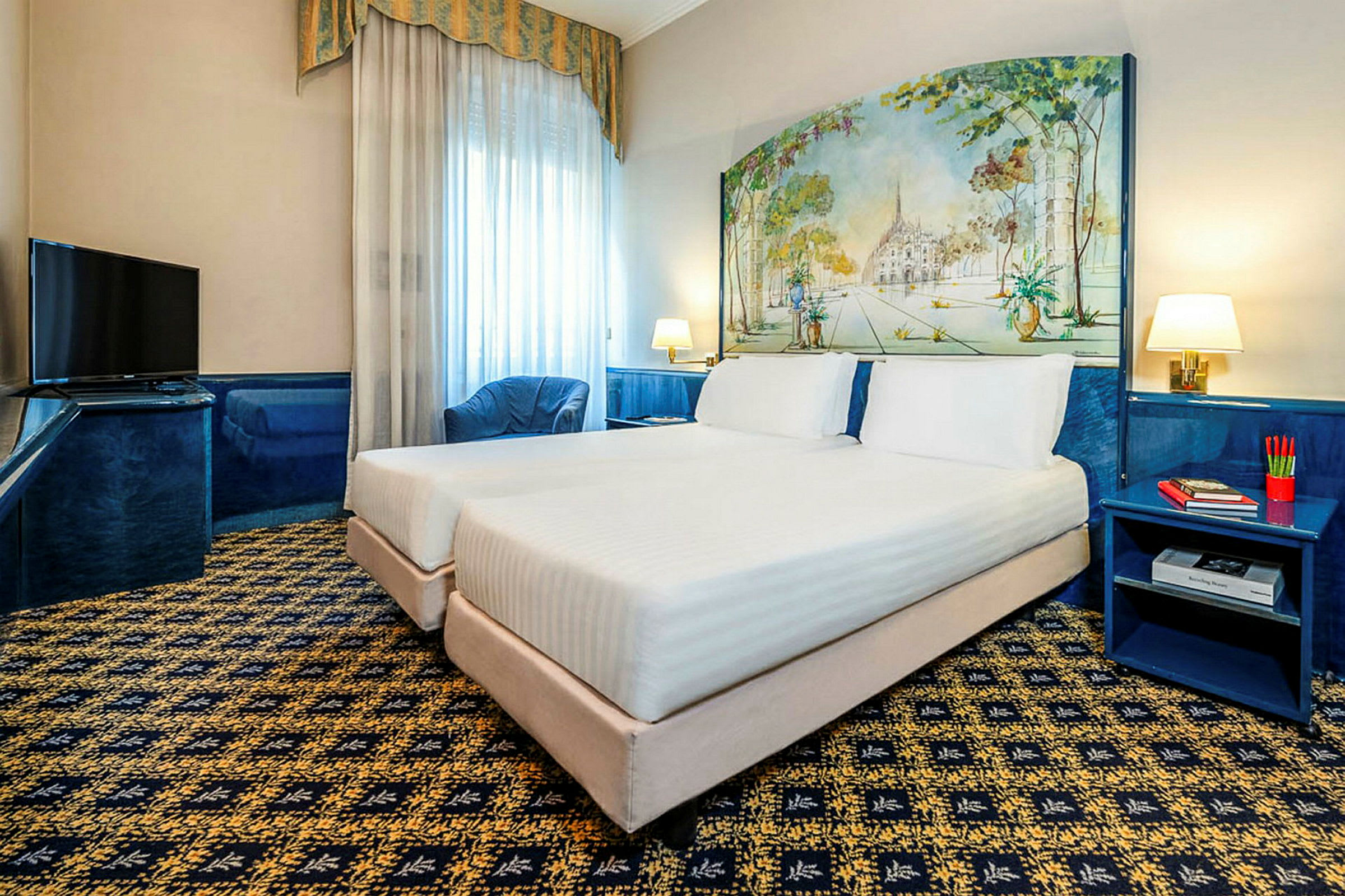 Ett hotellrum med två enkelsängar placerade intill varandra, ett mönstrat golv och ett målning som pryder väggen bakom sänggavlarna. Det finns en TV, en blå fåtölj och ett nattduksbord med böcker och pennor. (AI-genererad text)