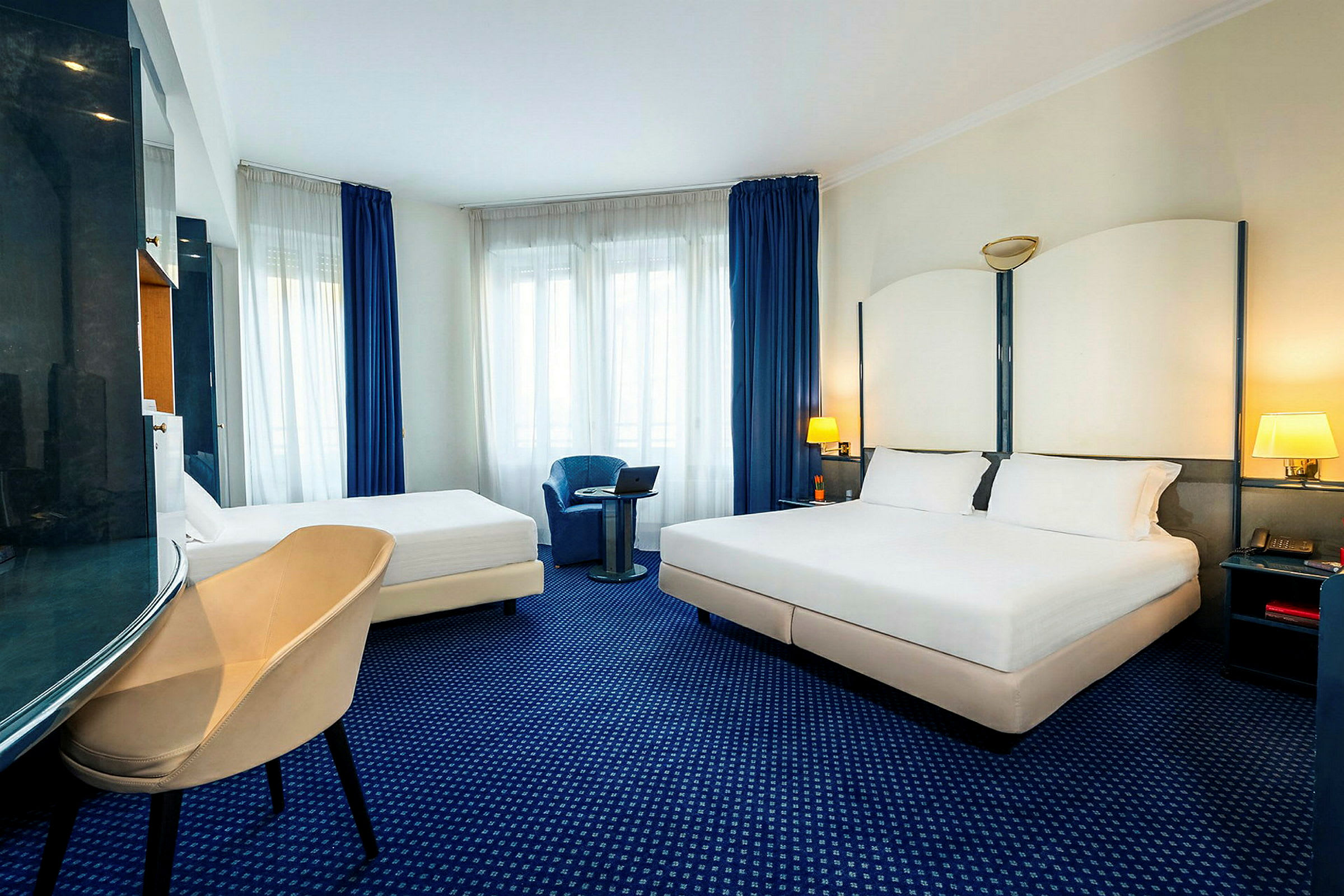 Hotellrum med en dubbelsäng och en enkelsäng, sittstol och fönster med blå gardiner. (AI-genererad text)