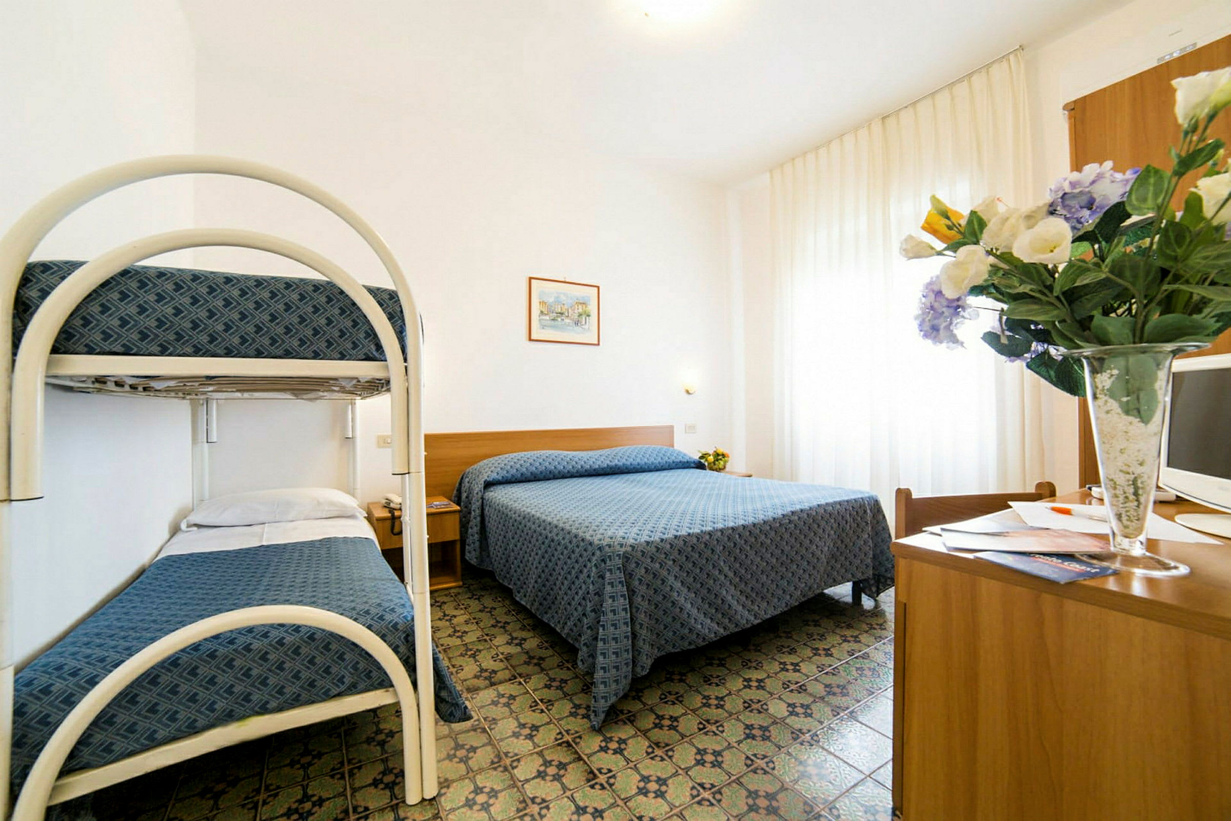 Ett hotellrum med en dubbelsäng och en våningssäng. På rummet finns även ett skrivbord med en TV och en vas med blommor. (AI-genererad text)