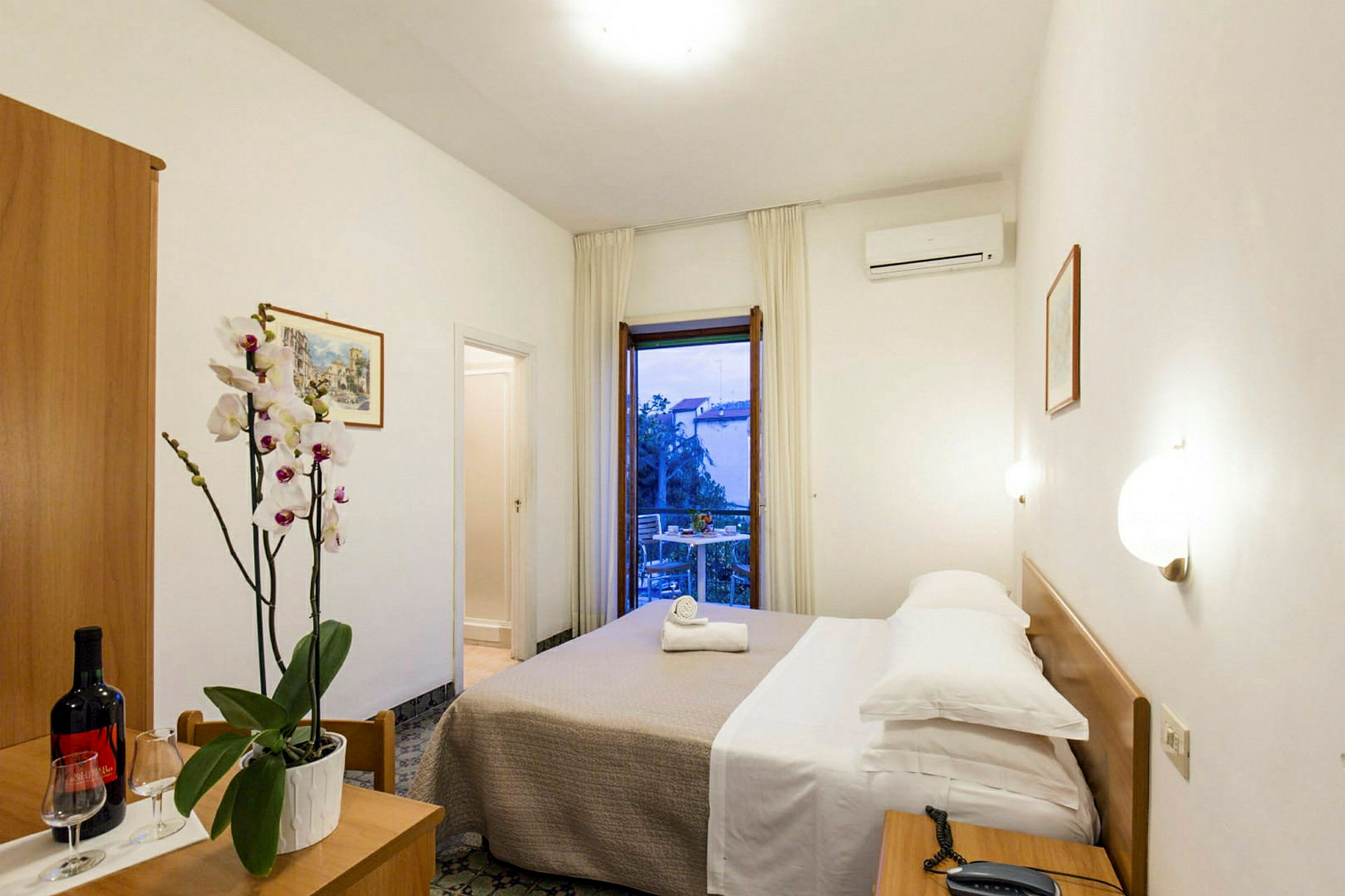 Ett hotellrum med en dubbelsäng, nattduksbord, en blomvas på ett litet bord och en balkong med utsikt. (AI-genererad text)