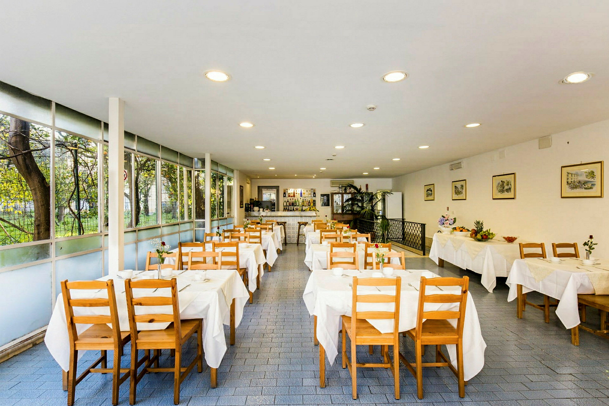 Ett ljust restaurangutrymme med trästolar och bord klädda i vita dukar. Fönster längs ena sidan ger utsikt mot grönska. (AI-genererad text)