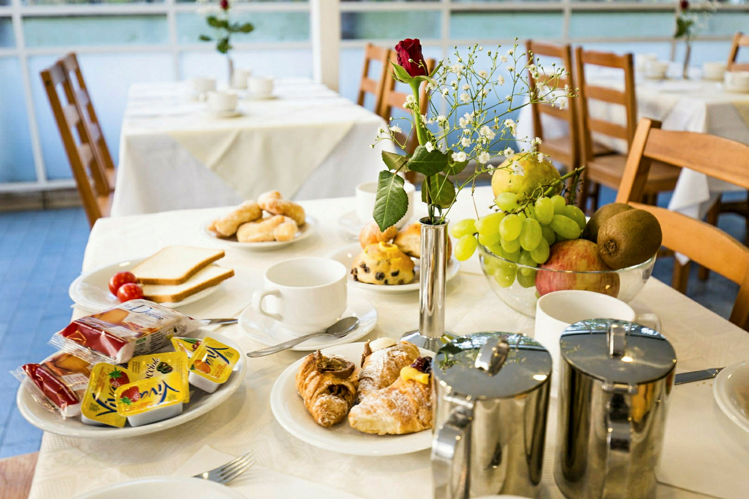 Frukostbord med vita dukar, fyllda med kaffe, juice, bröd, kakor, croissanter och frukt. En röd blomma står i en vas. (AI-genererad text)