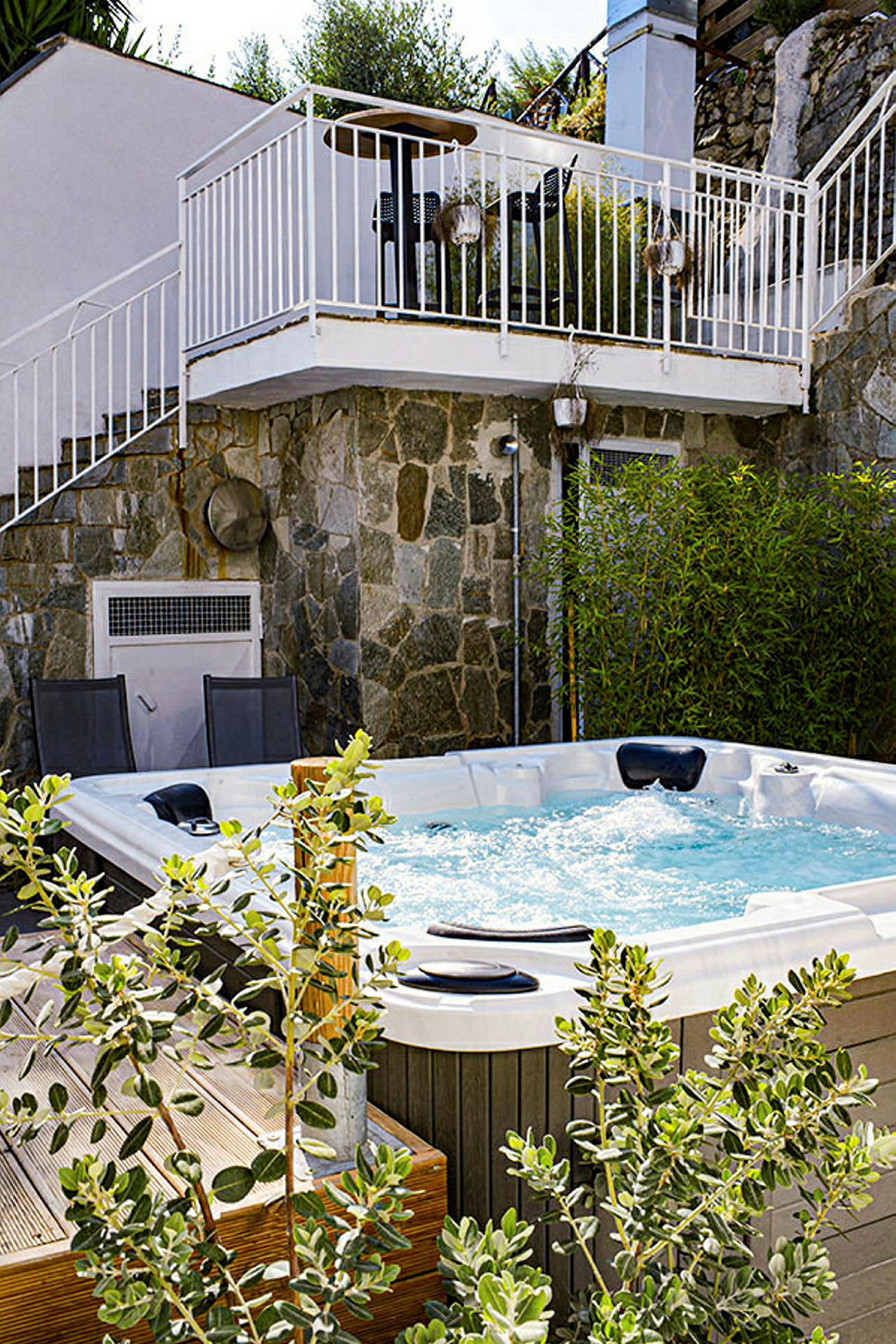 Ett utomhusbad med en jacuzzi omgiven av grönska. I bakgrunden syns en mur och en terrass. (AI-genererad text)