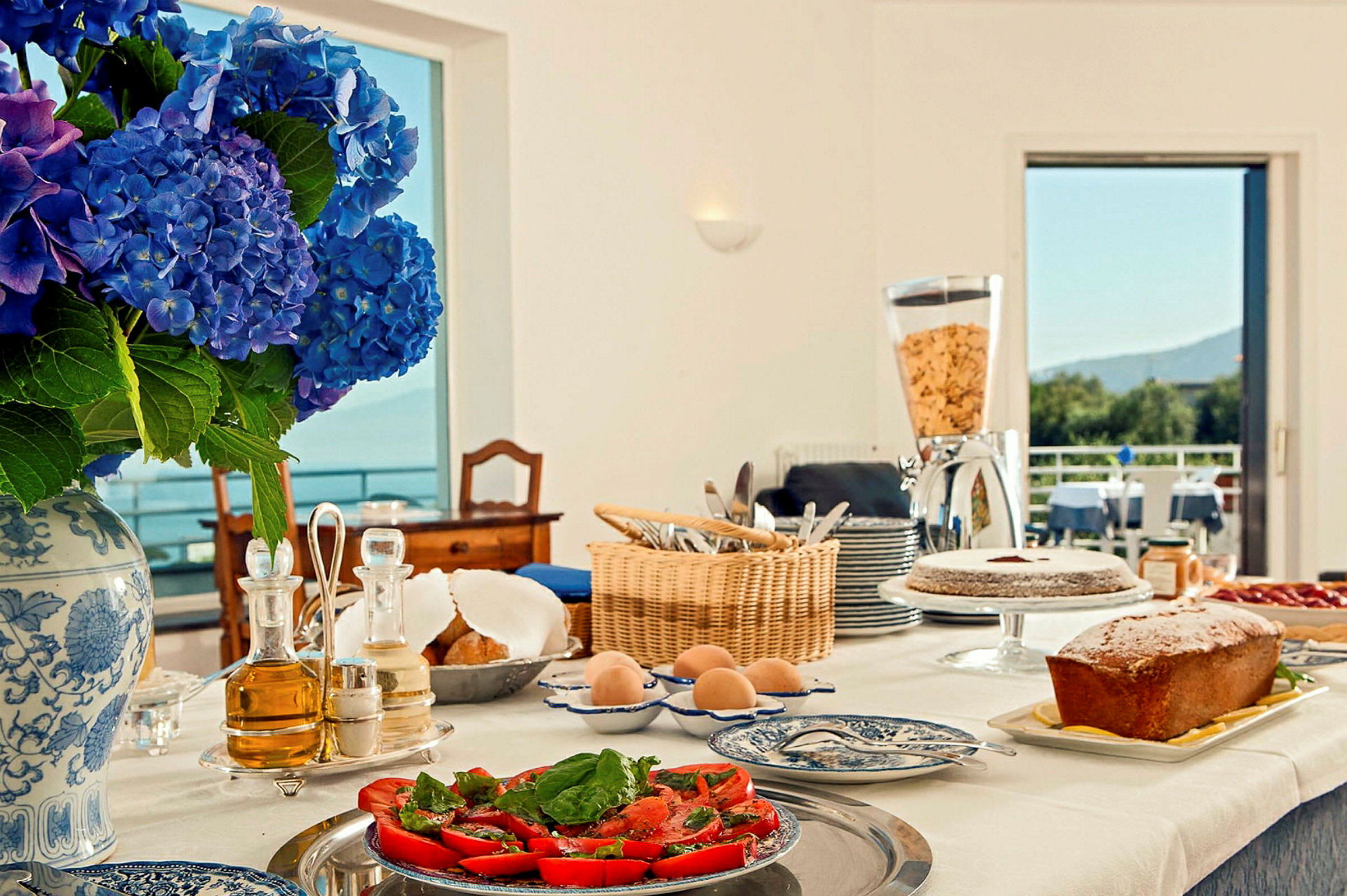 Ett bord med frukostmat dukat, inklusive en vas med blå hortensior, bröd, kakor, tomater och andra rätter, med en öppning till en balkong i bakgrunden. (AI-genererad text)