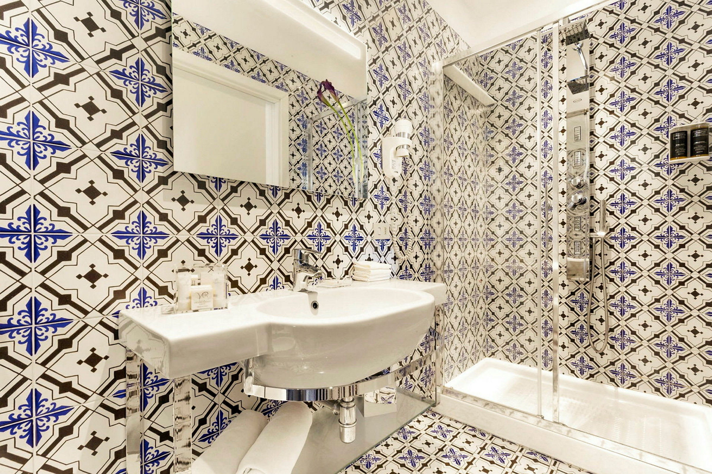 Ett badrum med mönstrade kakelplattor på väggar och golv, en vit handfat, och en glasomgiven dusch. (AI-genererad text)