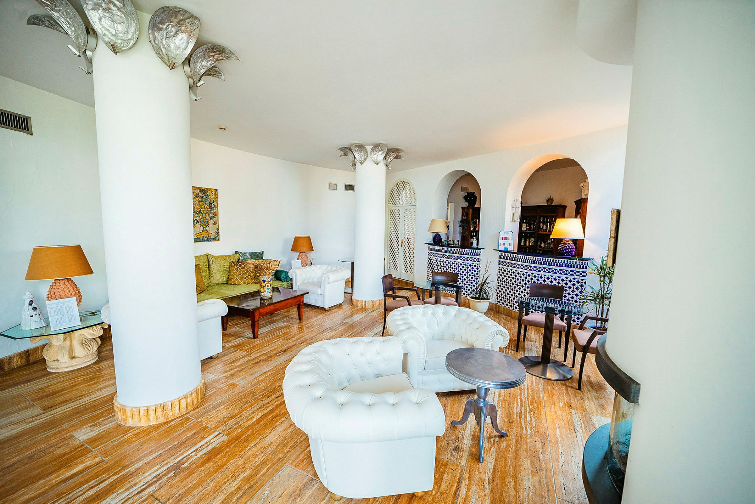 Ett vardagsrum med vita stoppade möbler, träbord och färgglada kuddar. Bakgrunden inkluderar dekorativa pelare och väggar med bågformade öppningar. (AI-genererad text)