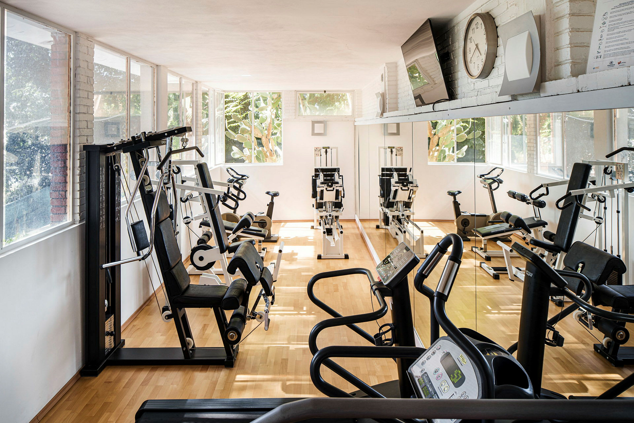 Ett gym med träningsmaskiner, inklusive stationära cyklar och löpband, på ett ljusinsläppande område. Väggarna har stora speglar och fönster. (AI-genererad text)
