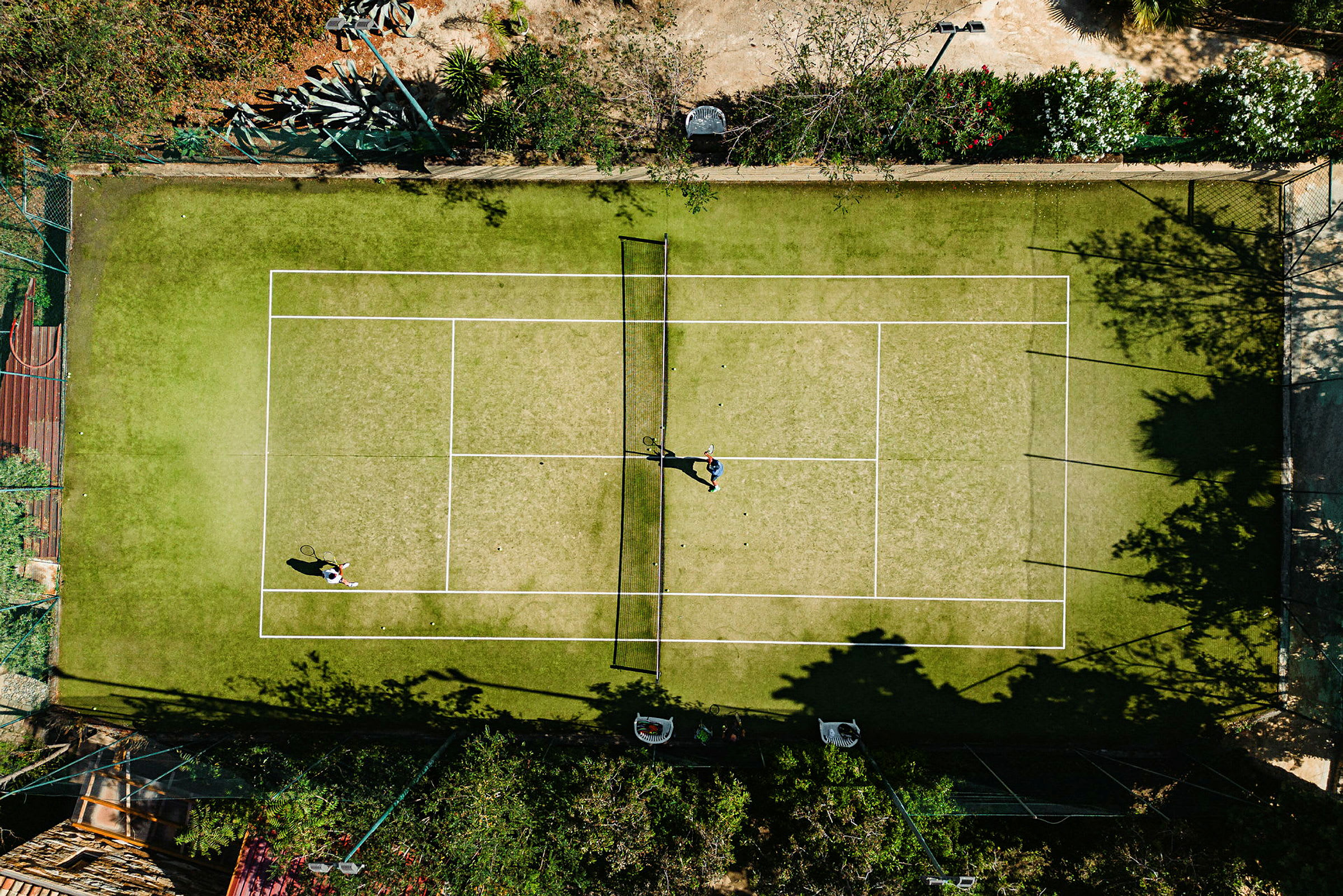 Flygfoto av två personer som spelar tennis på en grusplan omgivet av växtlighet. (AI-genererad text)
