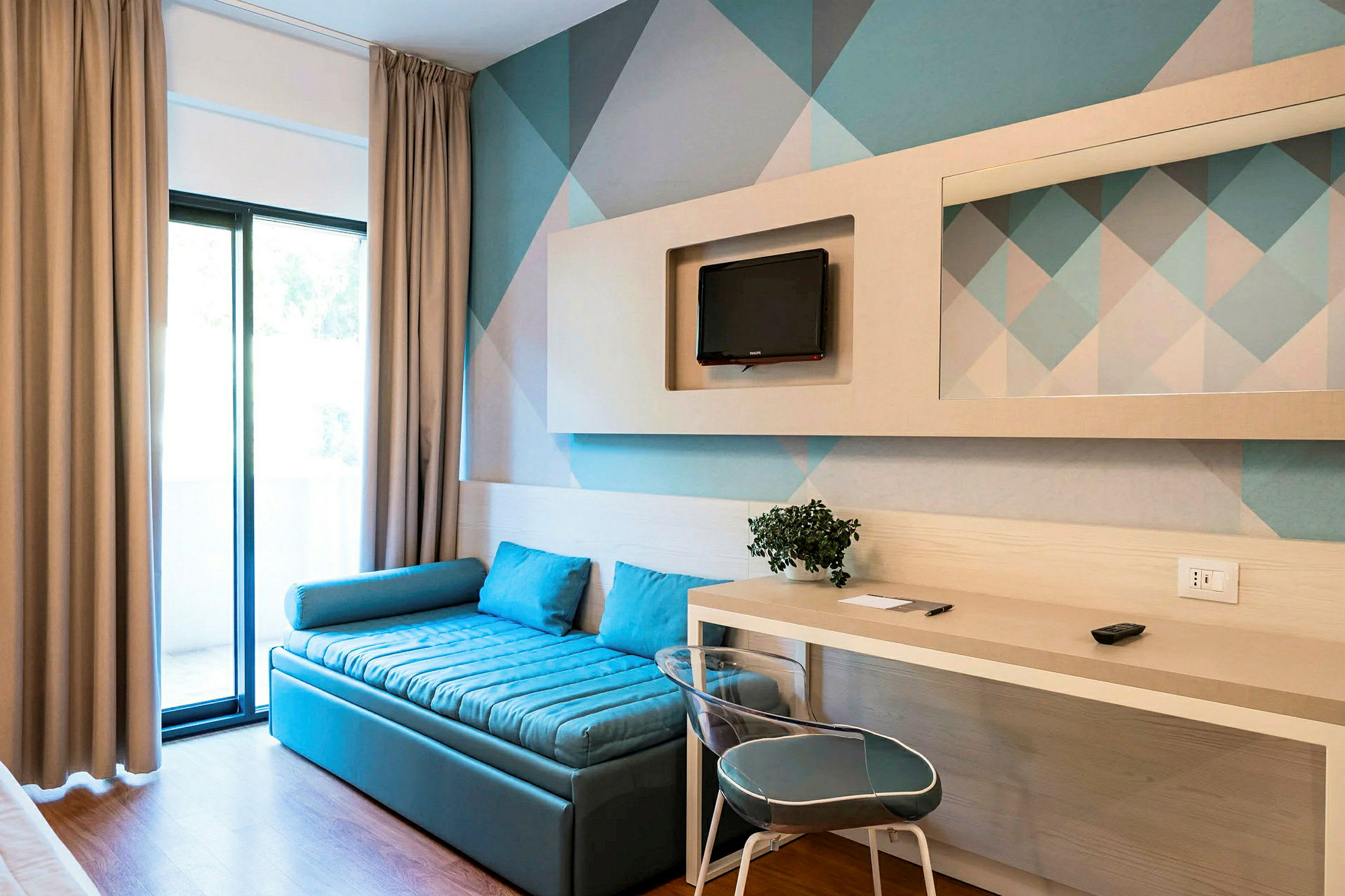 Ett hotellrum med en blå soffa, ett skrivbord, en stol och en väggmonterad TV. (AI-genererad text)