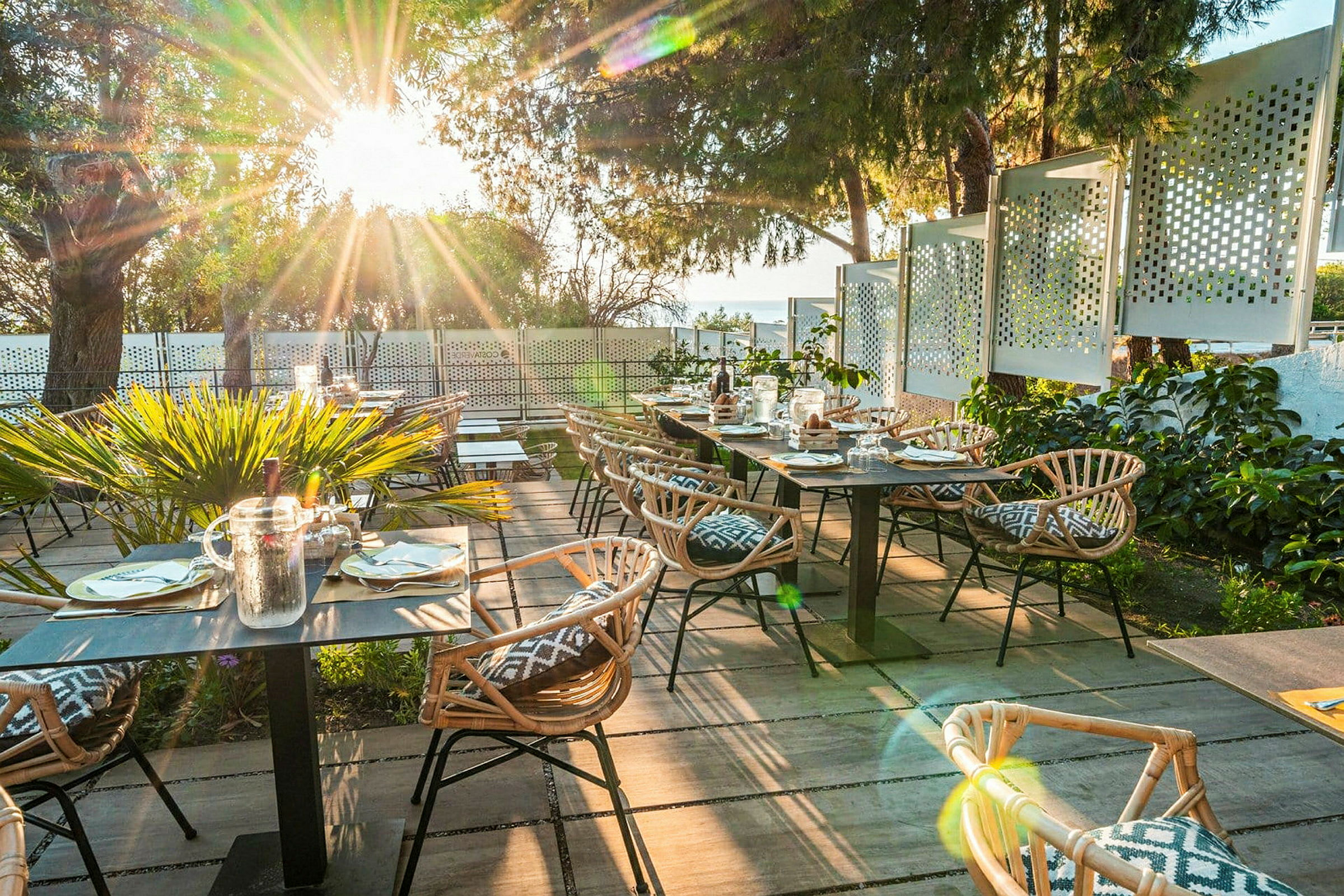 Utomhusrestaurang med träbord och rottingstolar samt solstrålar genom träden. (AI-genererad text)