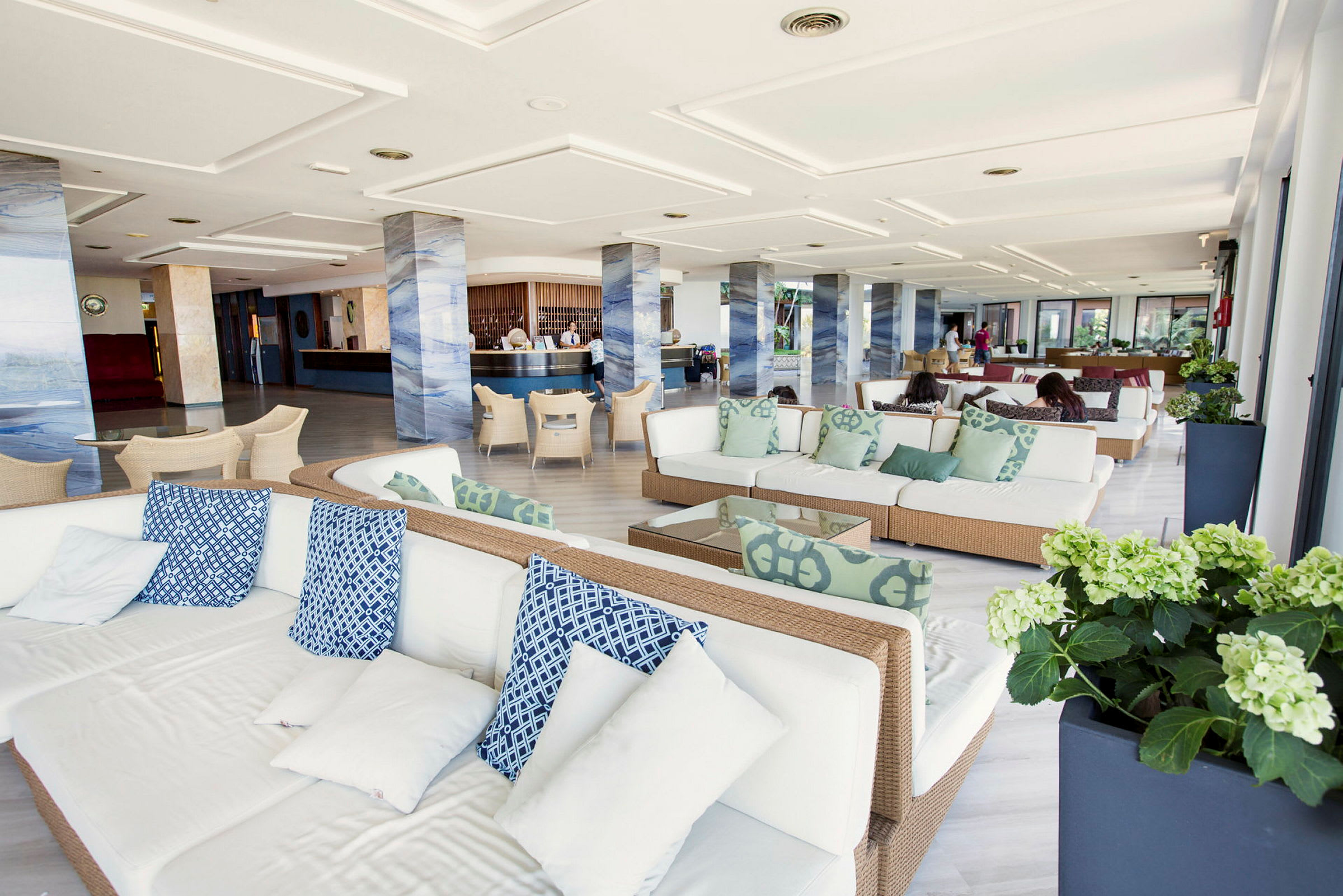 Ett modernt lobbyområde med vita soffor och blå samt gröna kuddar. Området har pelare i en marmorerad design och ett receptionsområde i bakgrunden. (AI-genererad text)