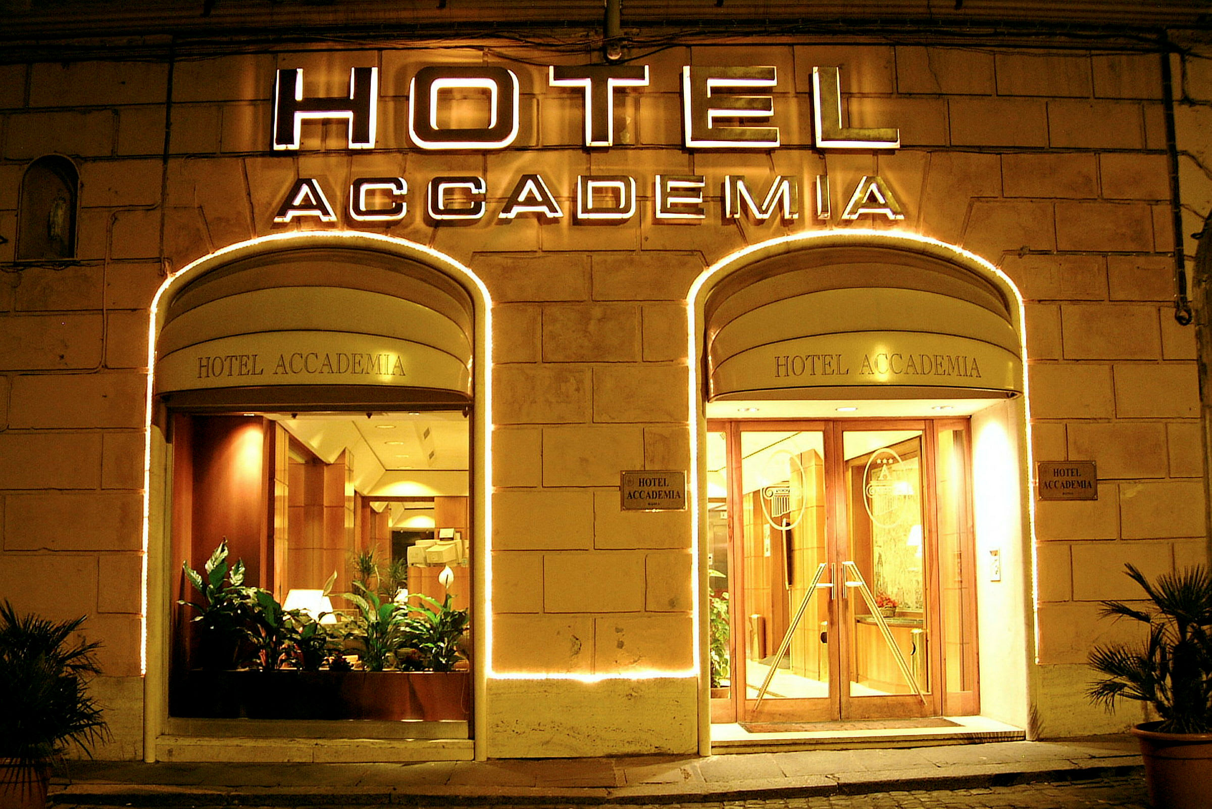 Hotell Accademias upplysta entré med glasdörrar och växter. (AI-genererad text)