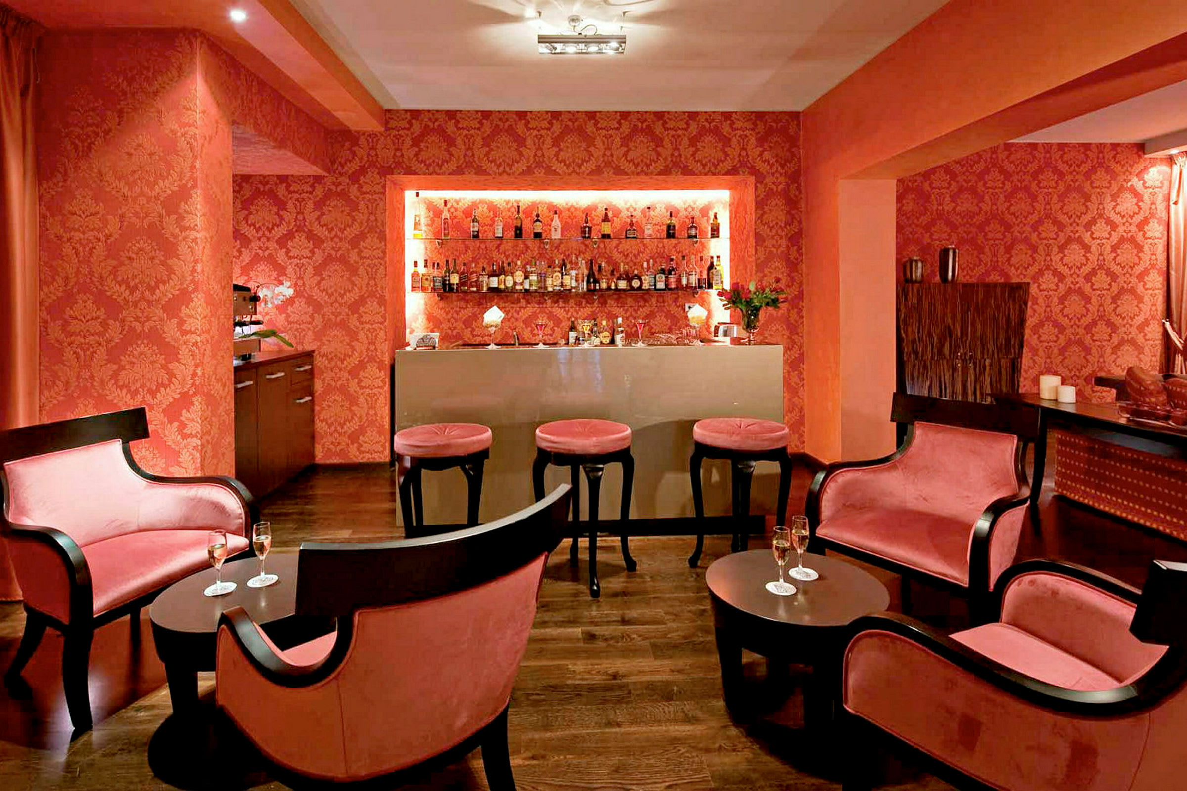 Bild som visar en elegant utrymme inrett som en cocktailbar med mörka möbler, rosa dynor och flaskor på en hylla. (AI-genererad text)