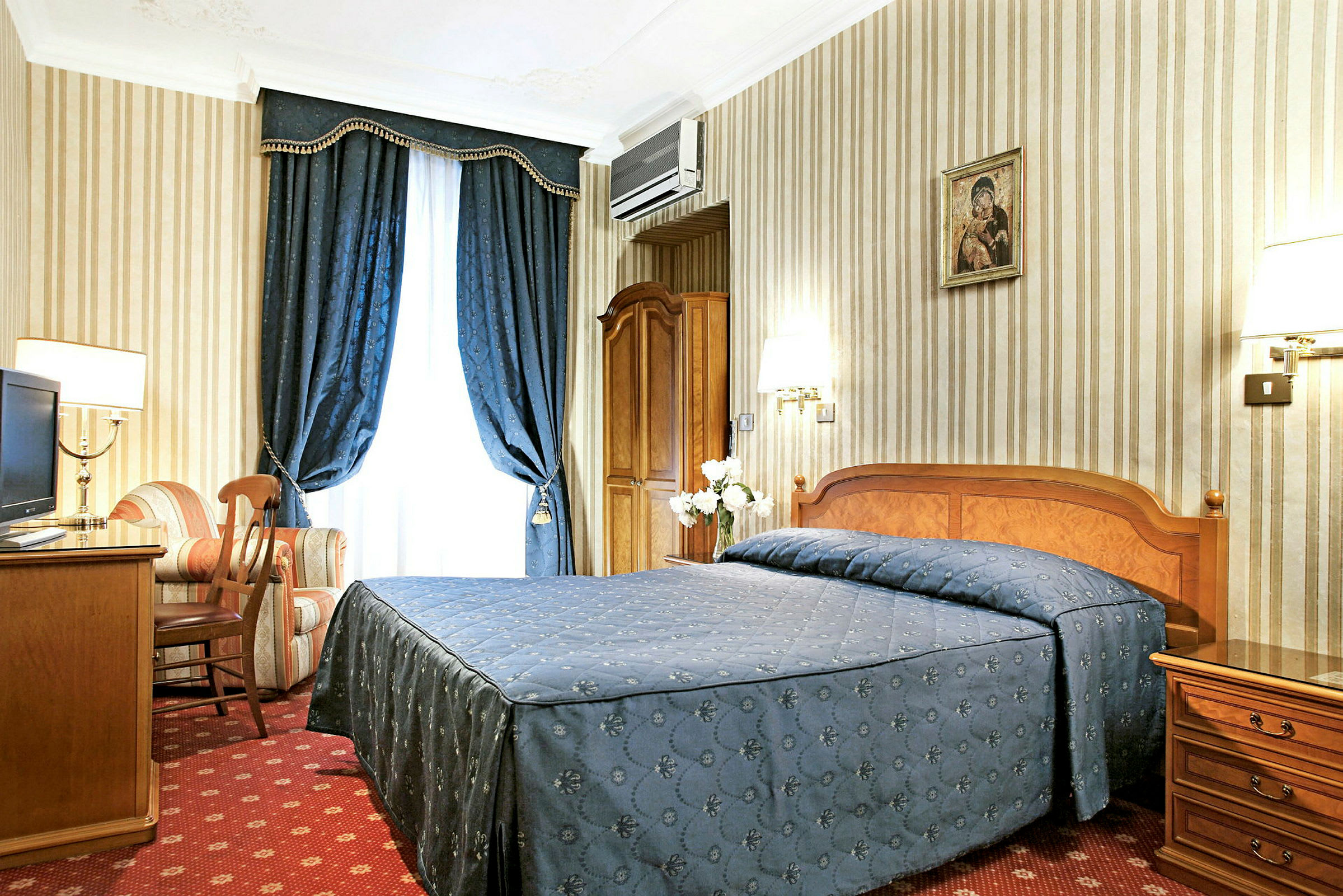 Ett hotellrum med en dubbelsäng klädd i blått överkast, ett skrivbord, en fåtölj och blå gardiner. (AI-genererad text)