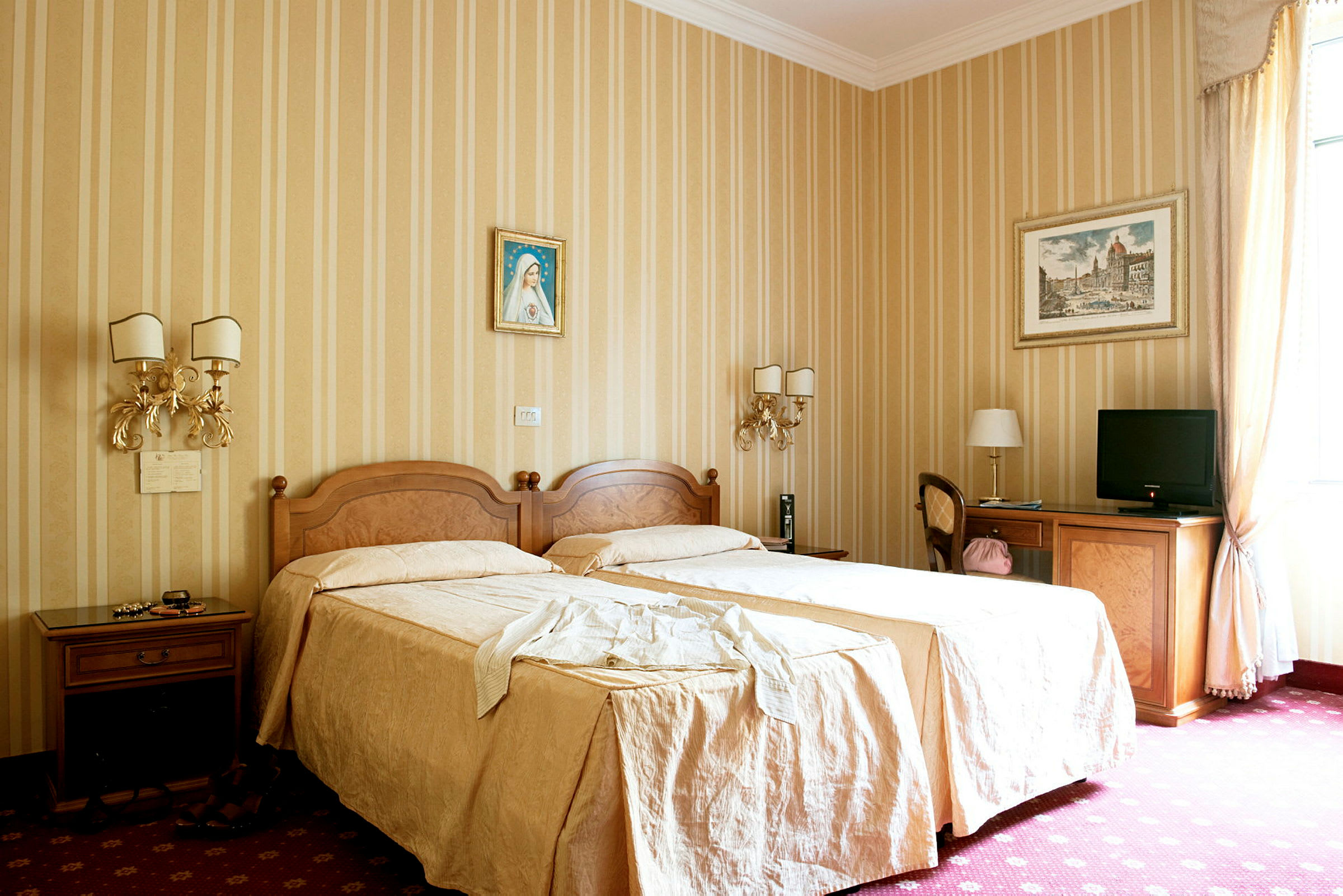 Ett hotellrum med två enkelsängar täckta med beige sängkläder, dekorerade väggar med tavlor, och möbler inklusive bord, stolar och en TV. (AI-genererad text)
