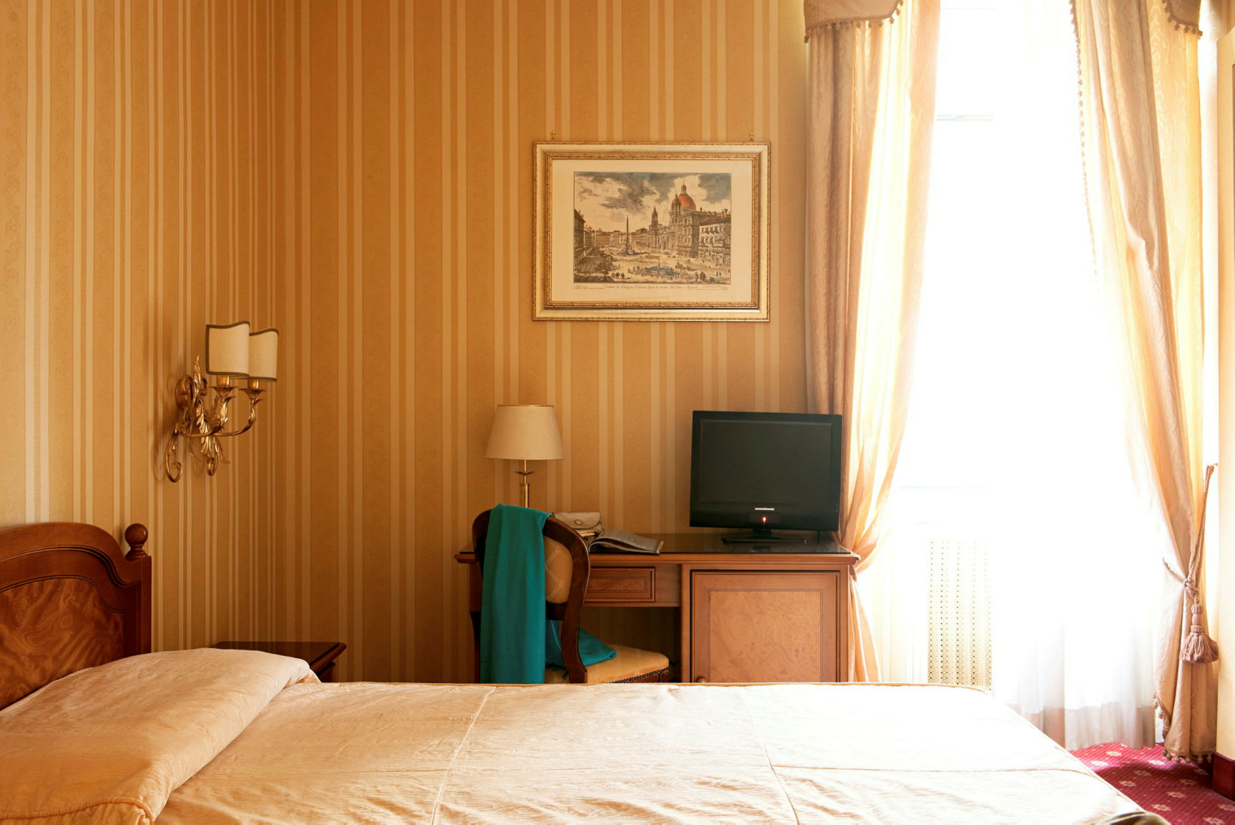 Ett hotellrum med en säng, nattduksbord, skrivbord med en stol och en TV, samt guldfärgade väggar och gardiner. (AI-genererad text)