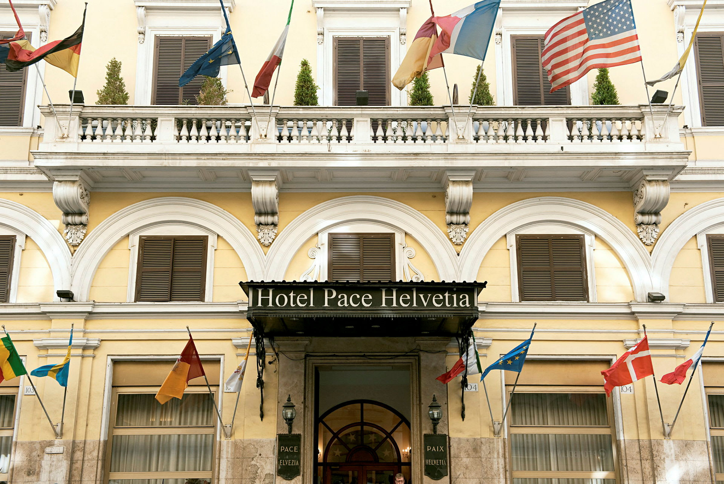 Hotellfasad med namnet 'Hotel Pace Helvetia', dekorerad med flera länders flaggor. (AI-genererad text)