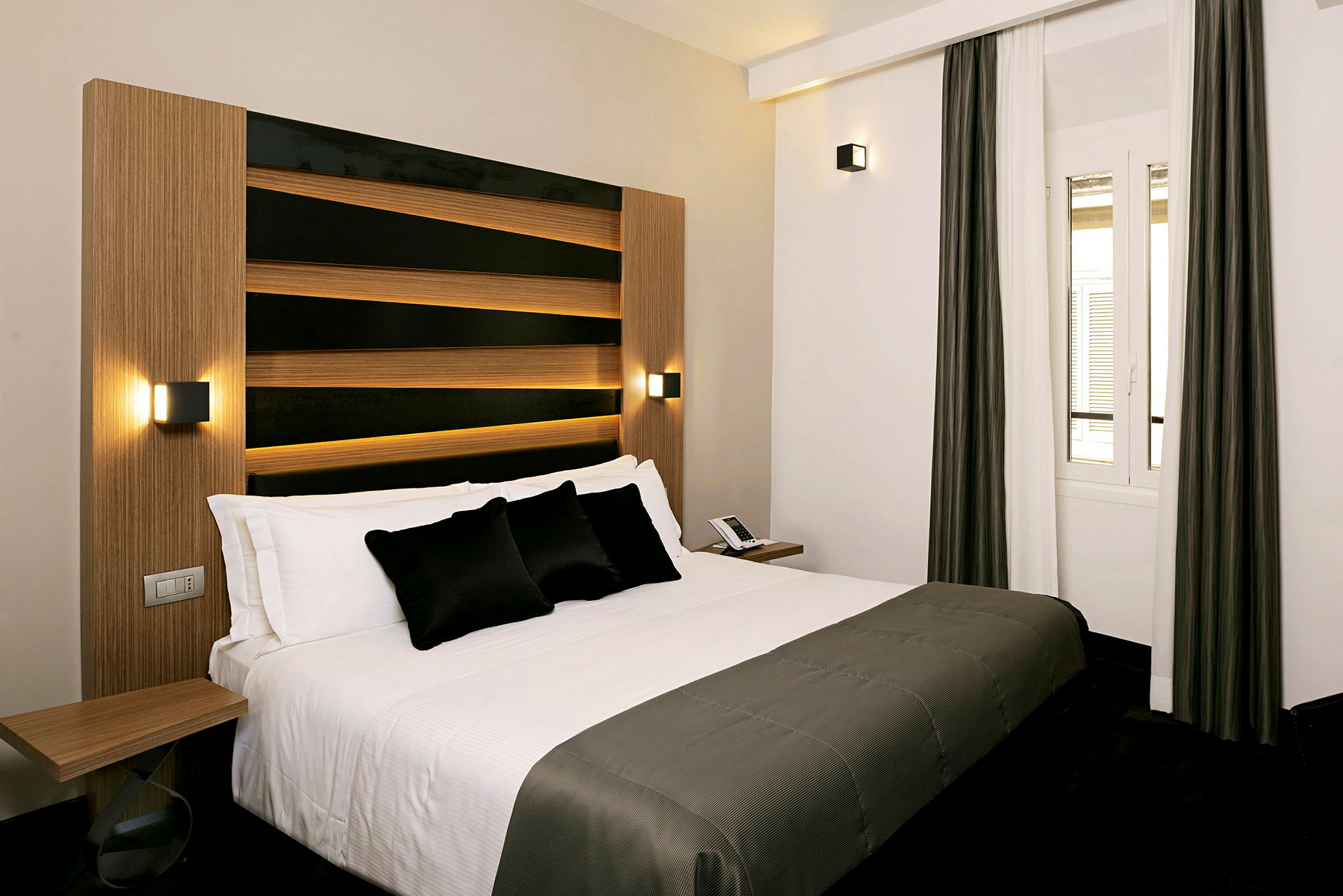 Denna bild visar ett hotellrum med en modern och elegant säng som har vita sängkläder och mörka dekorativa kuddar. Rummet har en enkel och stilren design med ett fönster med neutrala gardiner. (AI-genererad text)