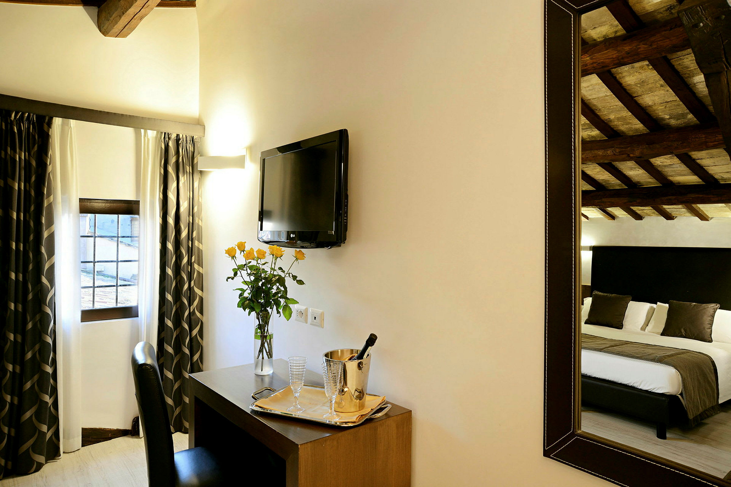 Ett hotellrum med draperier vid fönstret, en TV på väggen, ett skrivbord med en vas blommor och serveringsbricka samt en spegel som reflekterar en säng. (AI-genererad text)
