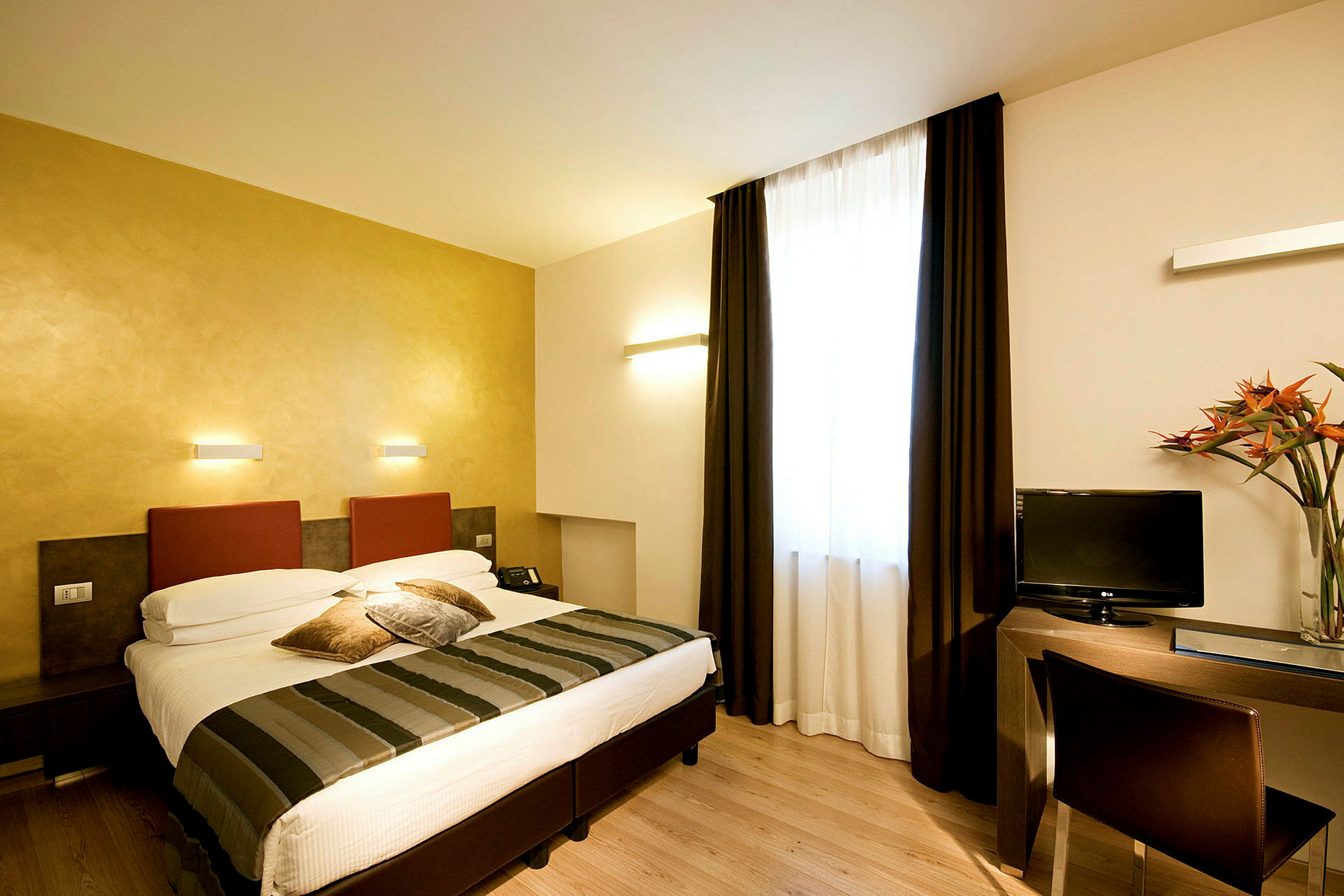Ett hotellrum med en dubbelsäng, skrivbord och TV. Rummet har ljusa väggar och ett stort fönster med mörka gardiner. (AI-genererad text)