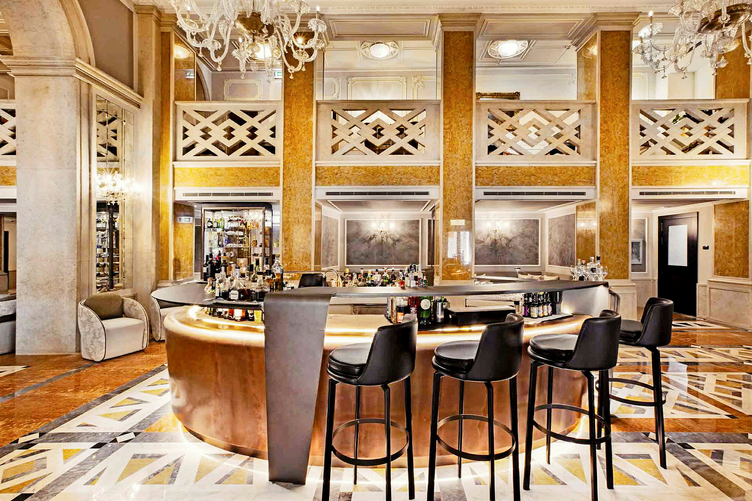 En elegant hotellbar, med polerade guldtonade material och en välvårdad golvyta, omringad av svarta barstolar och ståtliga takkronor. (AI-genererad text)