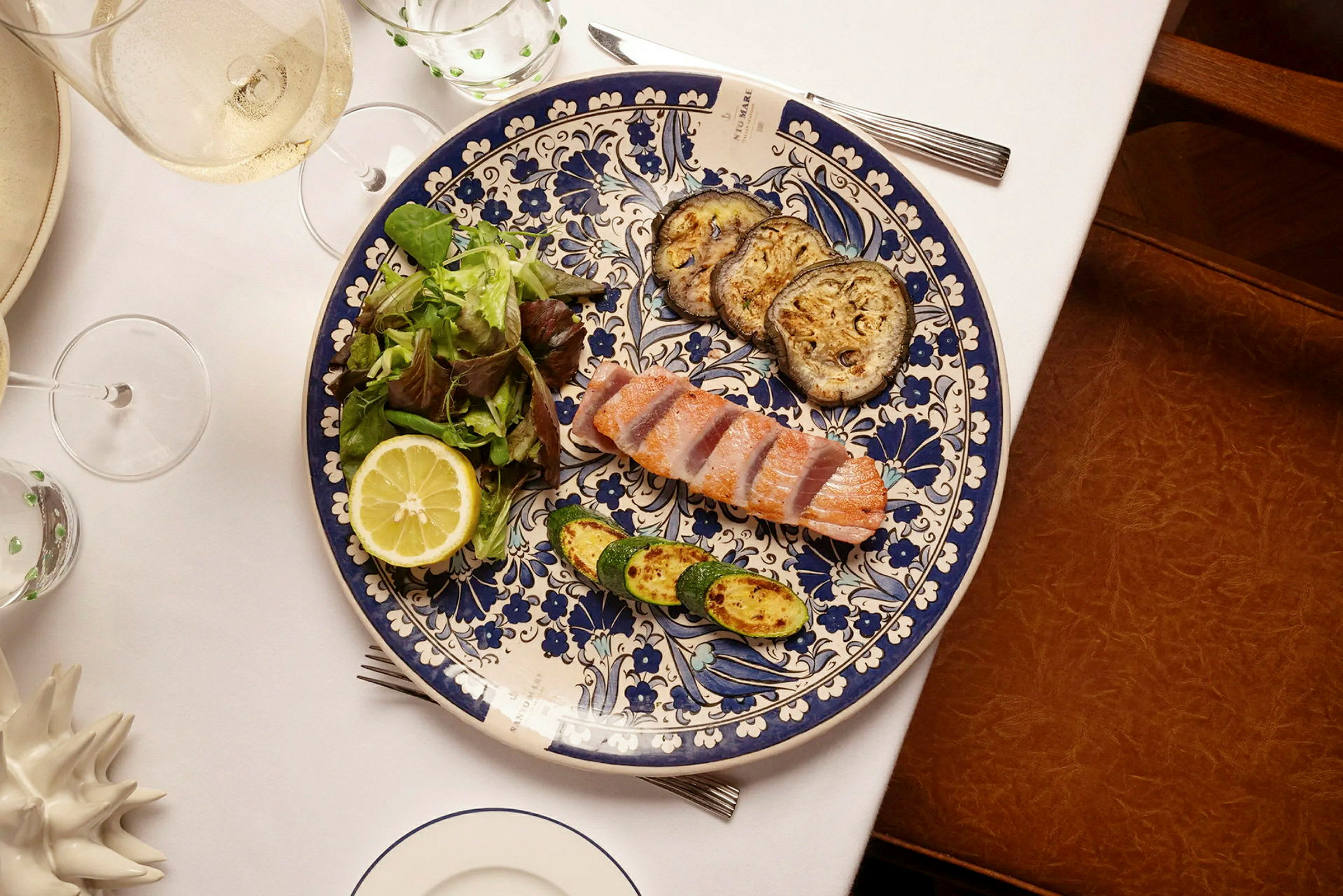 En tallrik innehållande bitar av lax, stekta grönsaker och sallad med en citronhalva, samt ett dukat bord. (AI-genererad text)